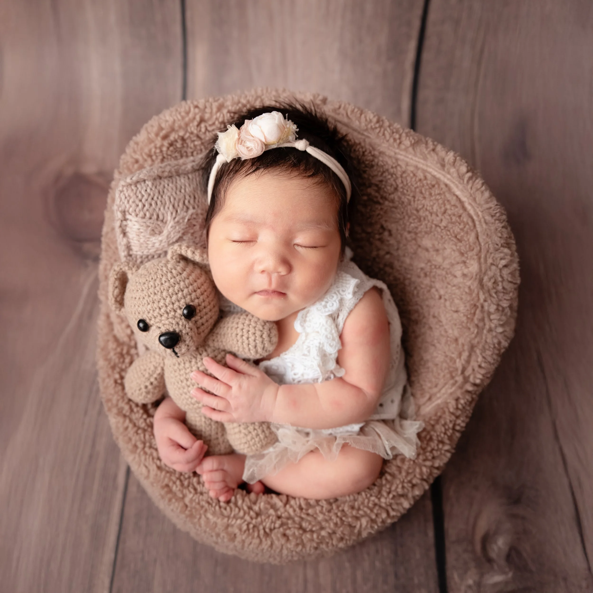Sleeping newborn wearing a pink floral headband curled in a soft brown basket while cuddling a small teddy bear