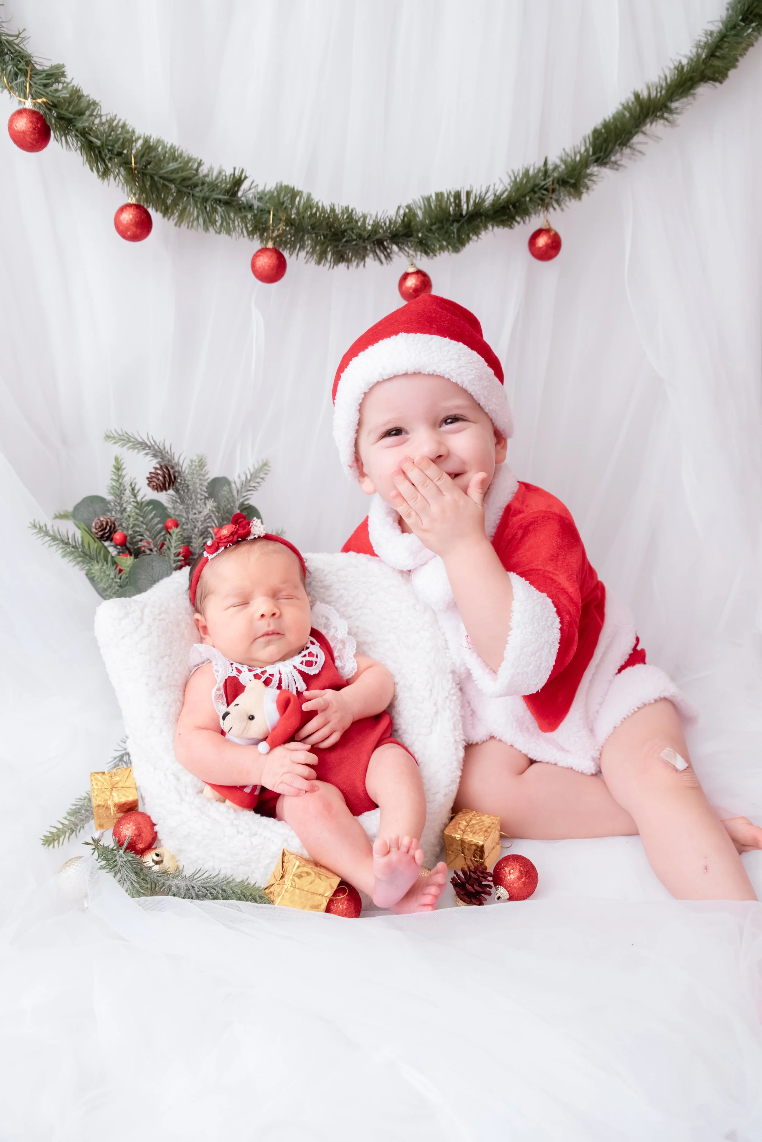 Smiling older sibling in a red Santa outfit sitting beside a sleeping newborn in a white basket beneath a Christmas garland and red baubles