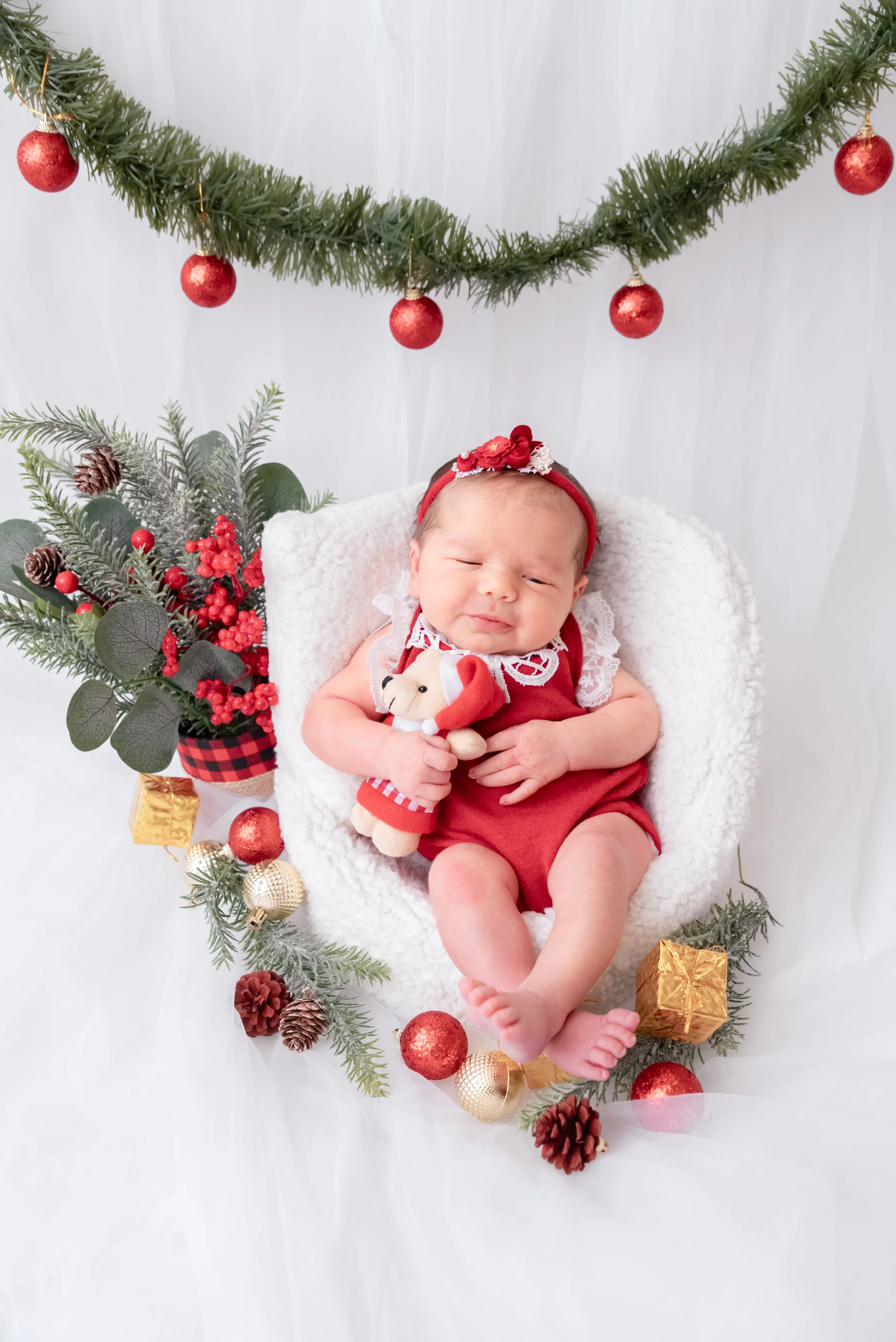 Sleeping newborn in a red festive outfit cuddling a teddy bear in a white basket beside Christmas greenery, red baubles and pine cones