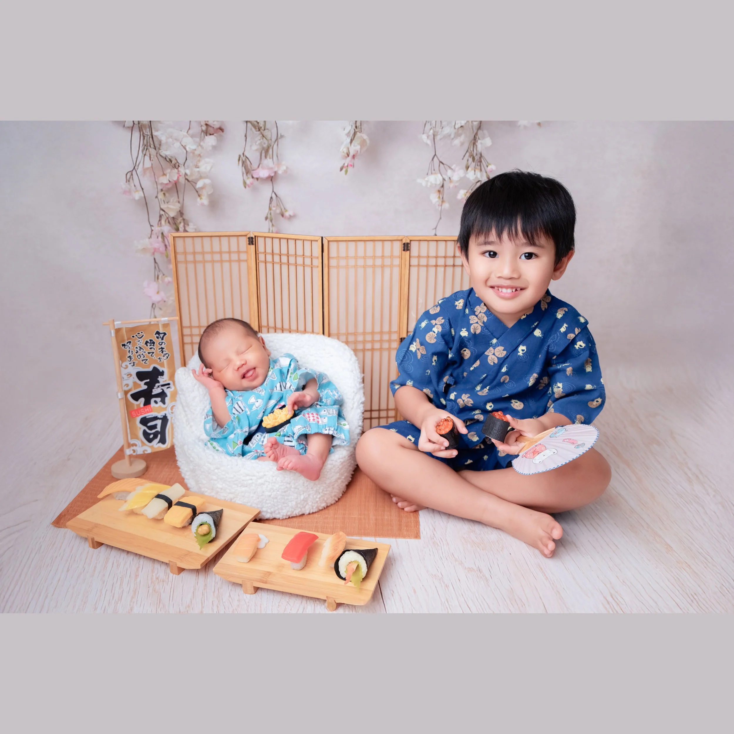Smiling older sibling in a navy patterned outfit sitting beside a sleeping newborn in a turquoise kimono in a white basket with toy sushi