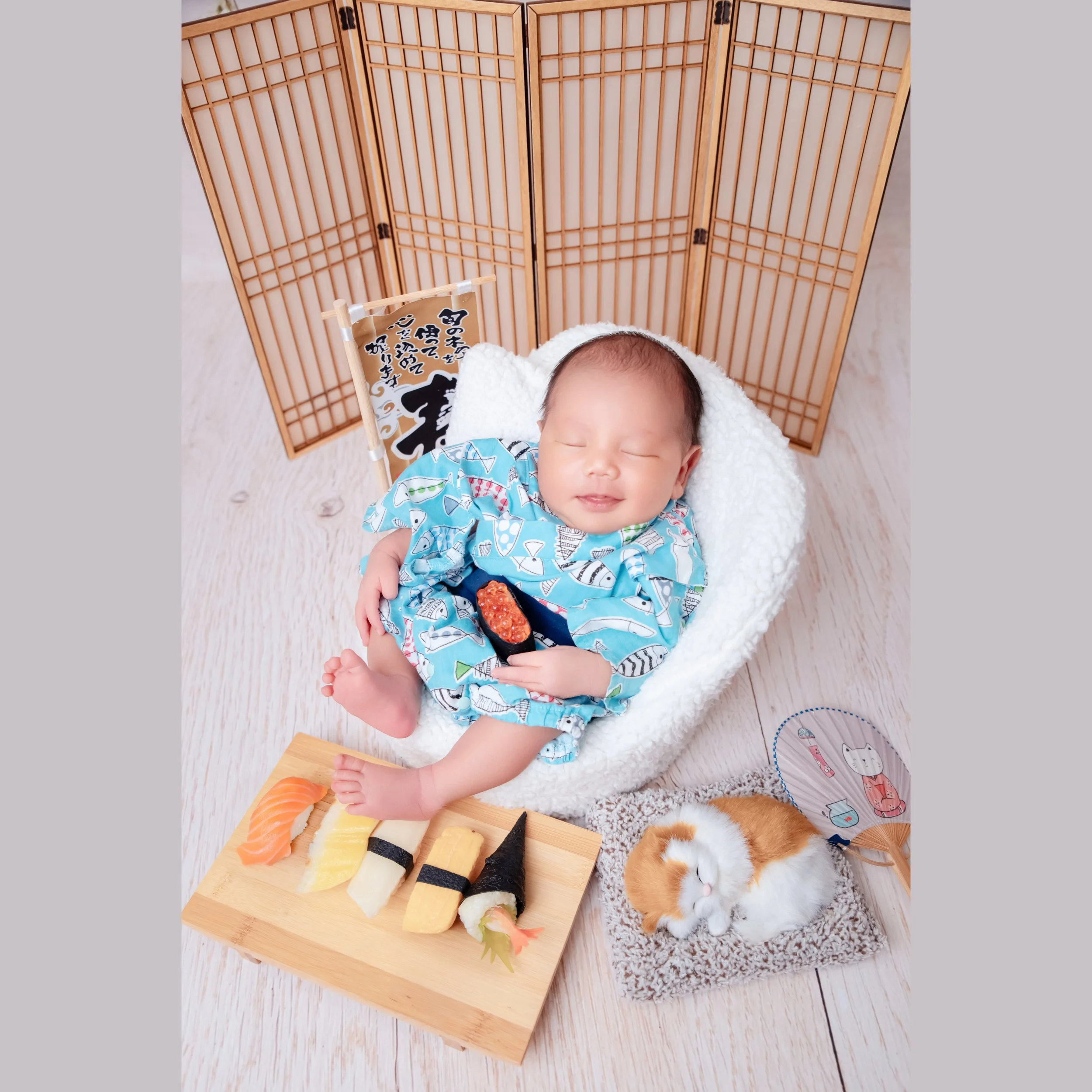 Sleeping newborn in a turquoise patterned kimono reclining in a white basket beside toy sushi and a wooden Japanese screen