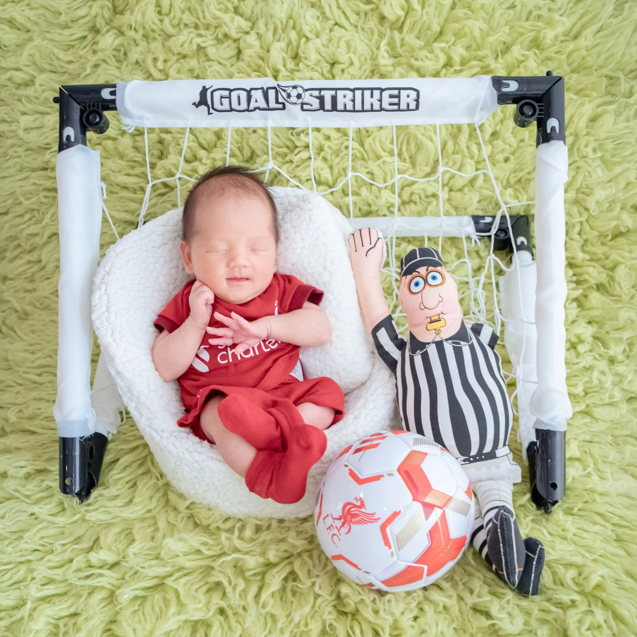 Newborn in a red Liverpool football kit reclining in a white basket inside a miniature goal beside a football and referee plush toy