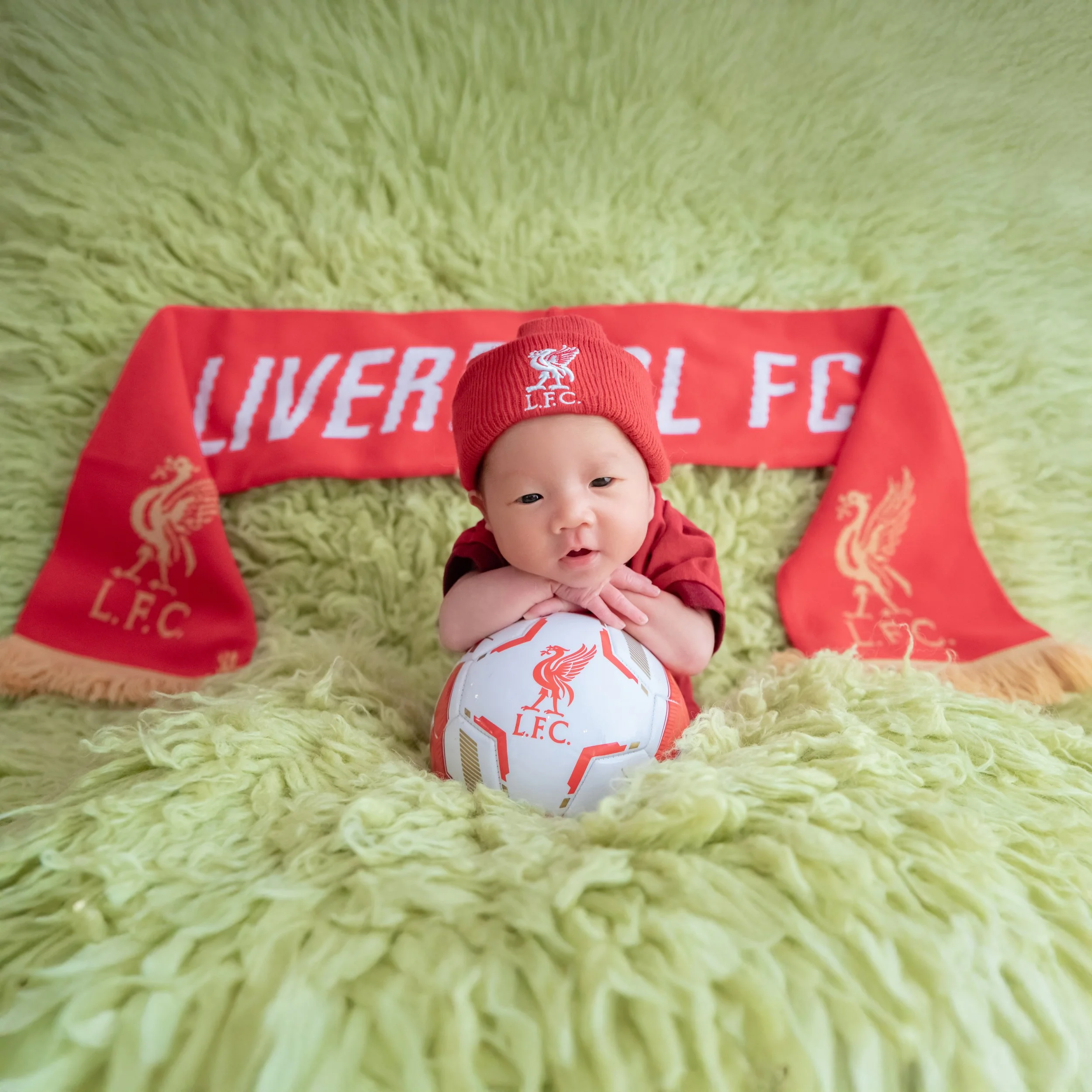 Awake newborn in a red Liverpool cap and outfit resting the chin on folded arms above a football on a green blanket with a Liverpool scarf behind