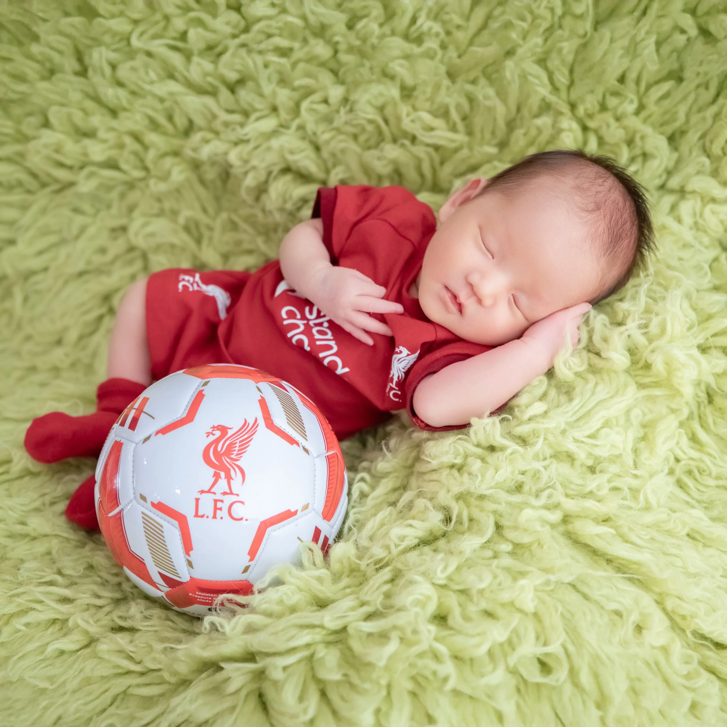 Sleeping newborn in a red Liverpool football outfit lying on one side on a green blanket with one hand beside the cheek and a football nearby
