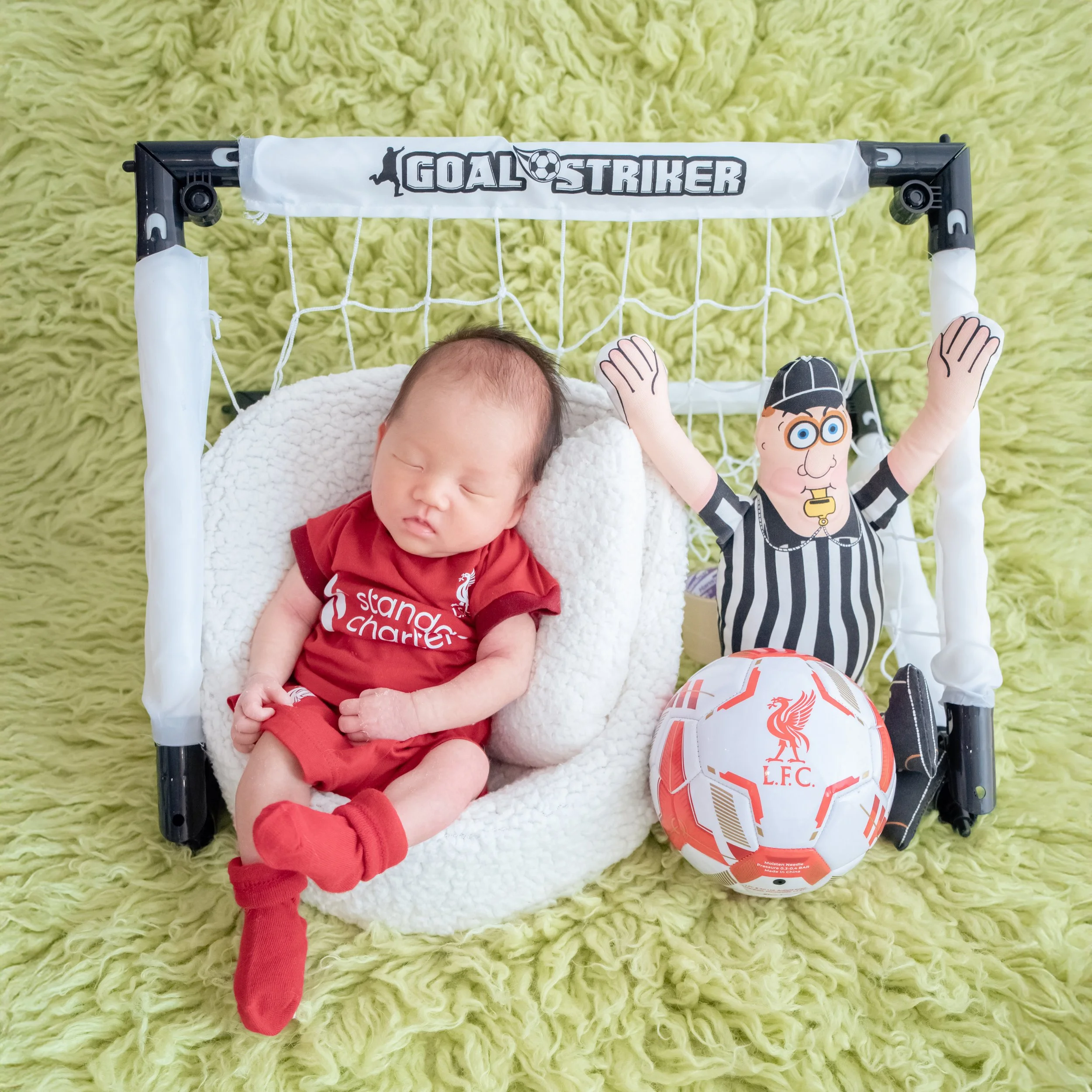 Sleeping newborn in a red Liverpool football kit reclining in a white basket inside a miniature goal beside a football and referee plush toy with raised arms