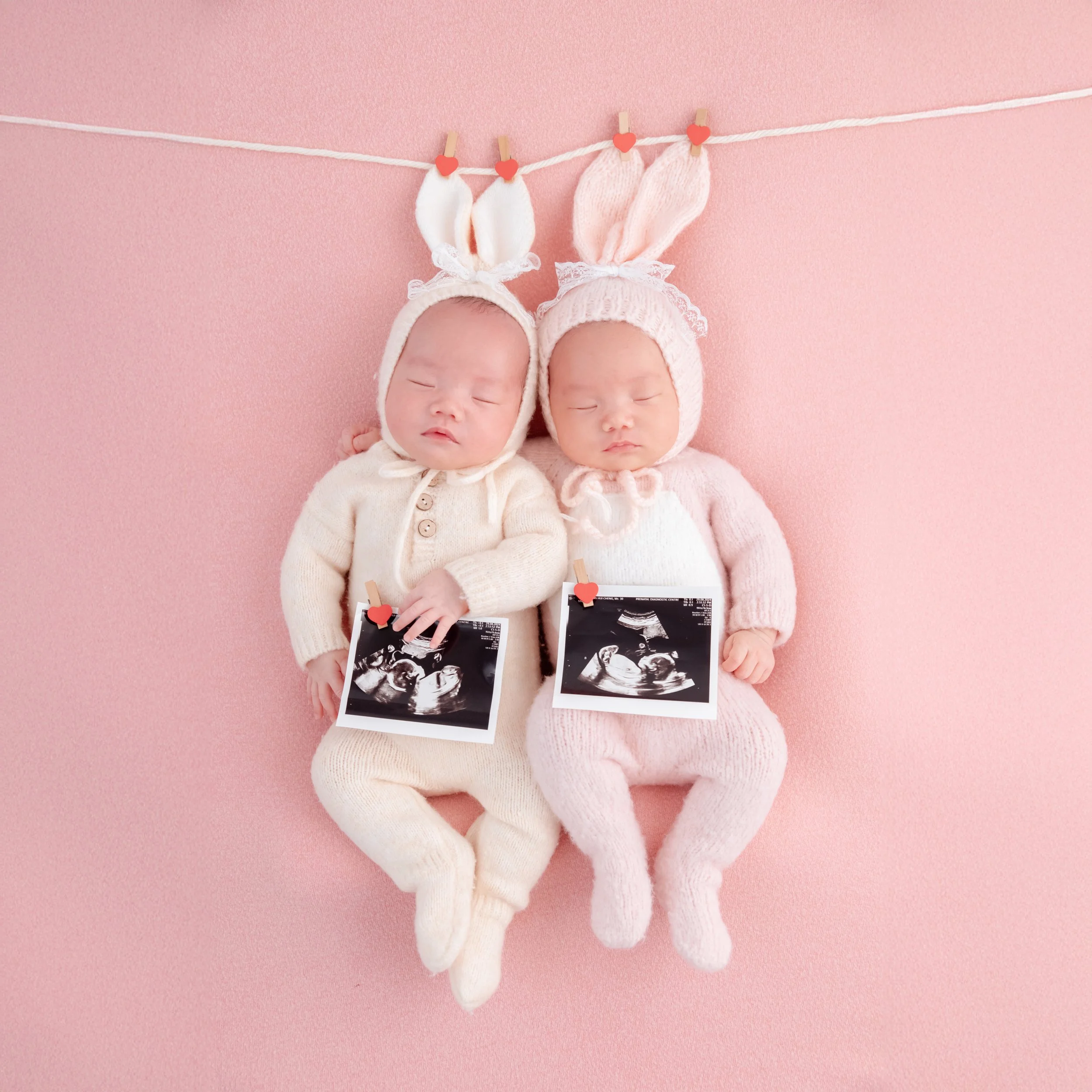Sleeping newborn twins in pink and cream knitted bunny outfits holding ultrasound prints on a pink backdrop