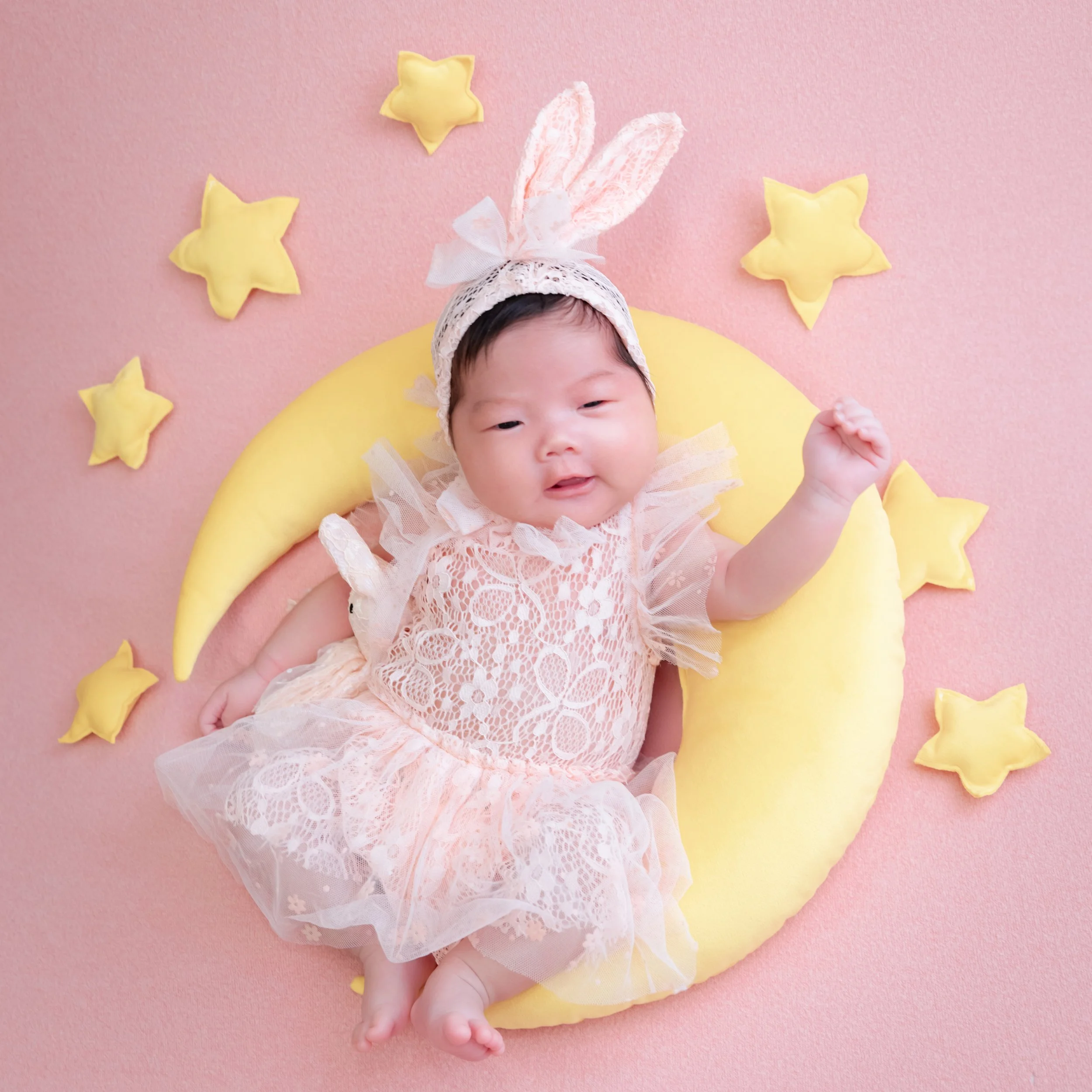 Newborn in a lace outfit and bunny ears lying on a yellow crescent moon with stars