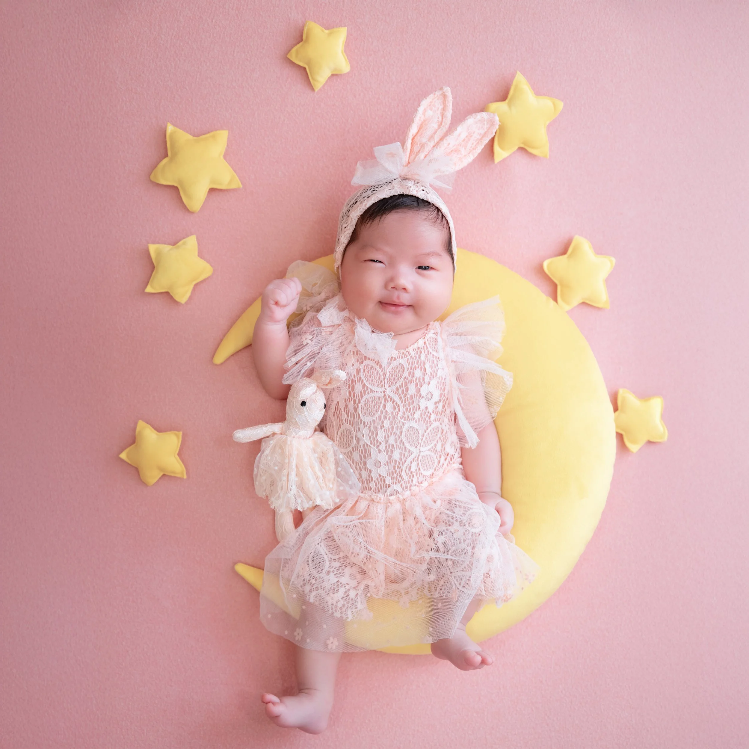 Smiling newborn in bunny ears holding a toy rabbit on a yellow moon against a pink backdrop