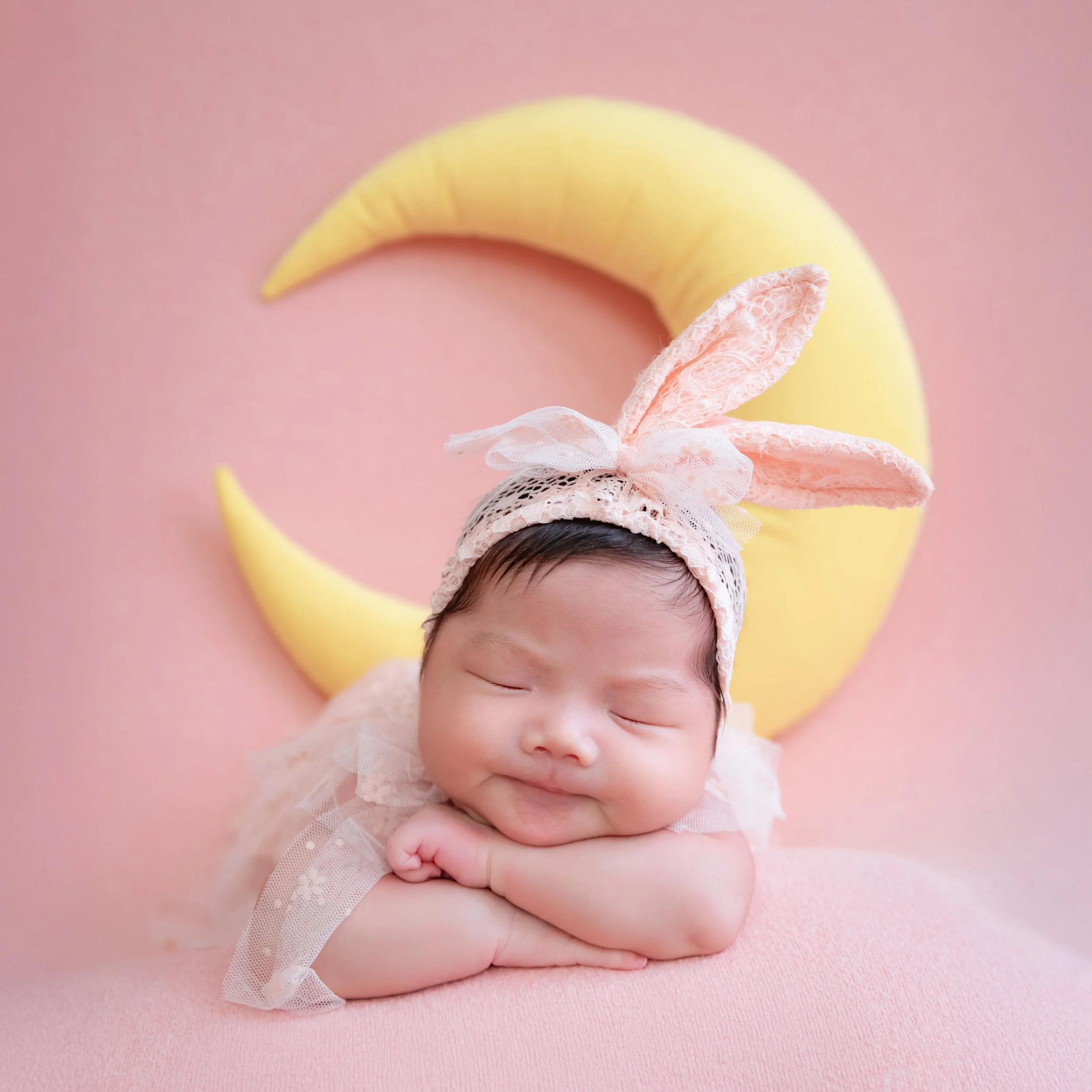 Sleeping newborn in bunny ears resting on folded hands with a yellow moon behind