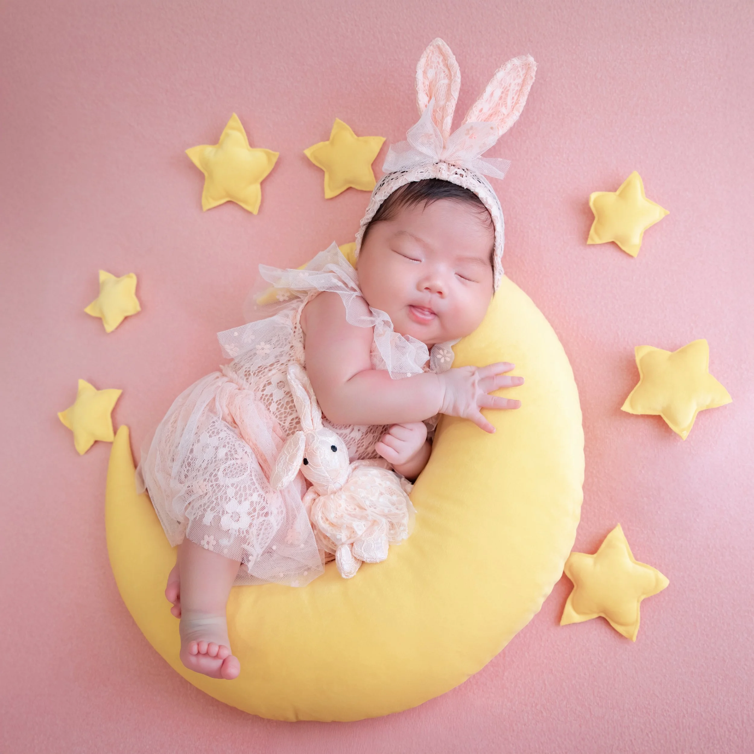 Sleeping newborn in bunny ears curled on a yellow crescent moon surrounded by stars