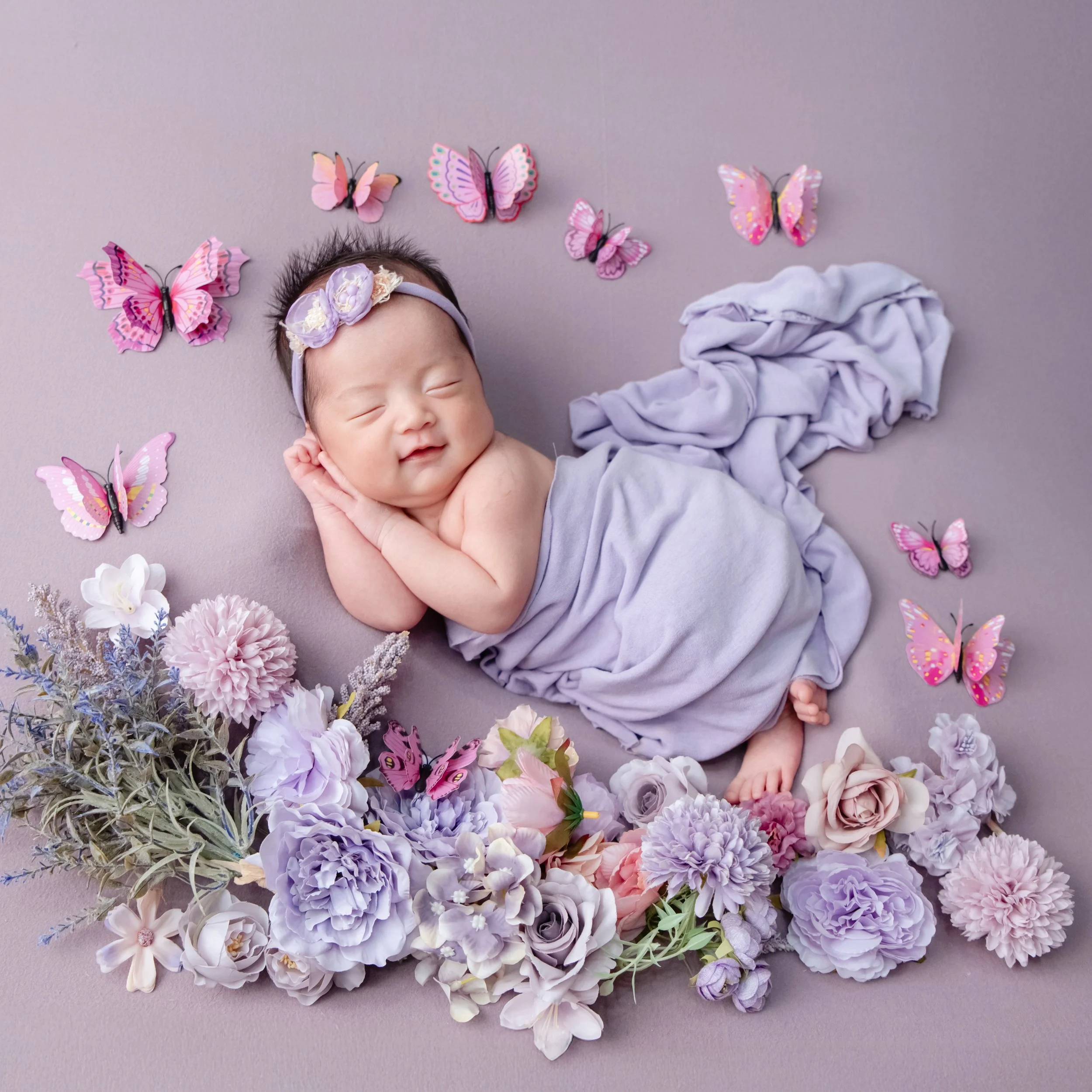 Smiling newborn wrapped in lavender fabric among pastel flowers and pink butterflies against a purple backdrop