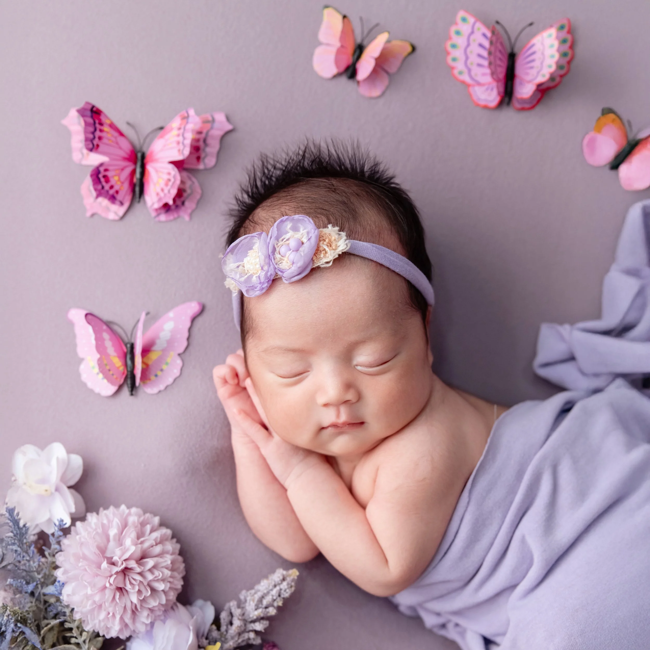 Sleeping newborn with hands tucked under one cheek, wrapped in lavender fabric among pink butterflies and flowers