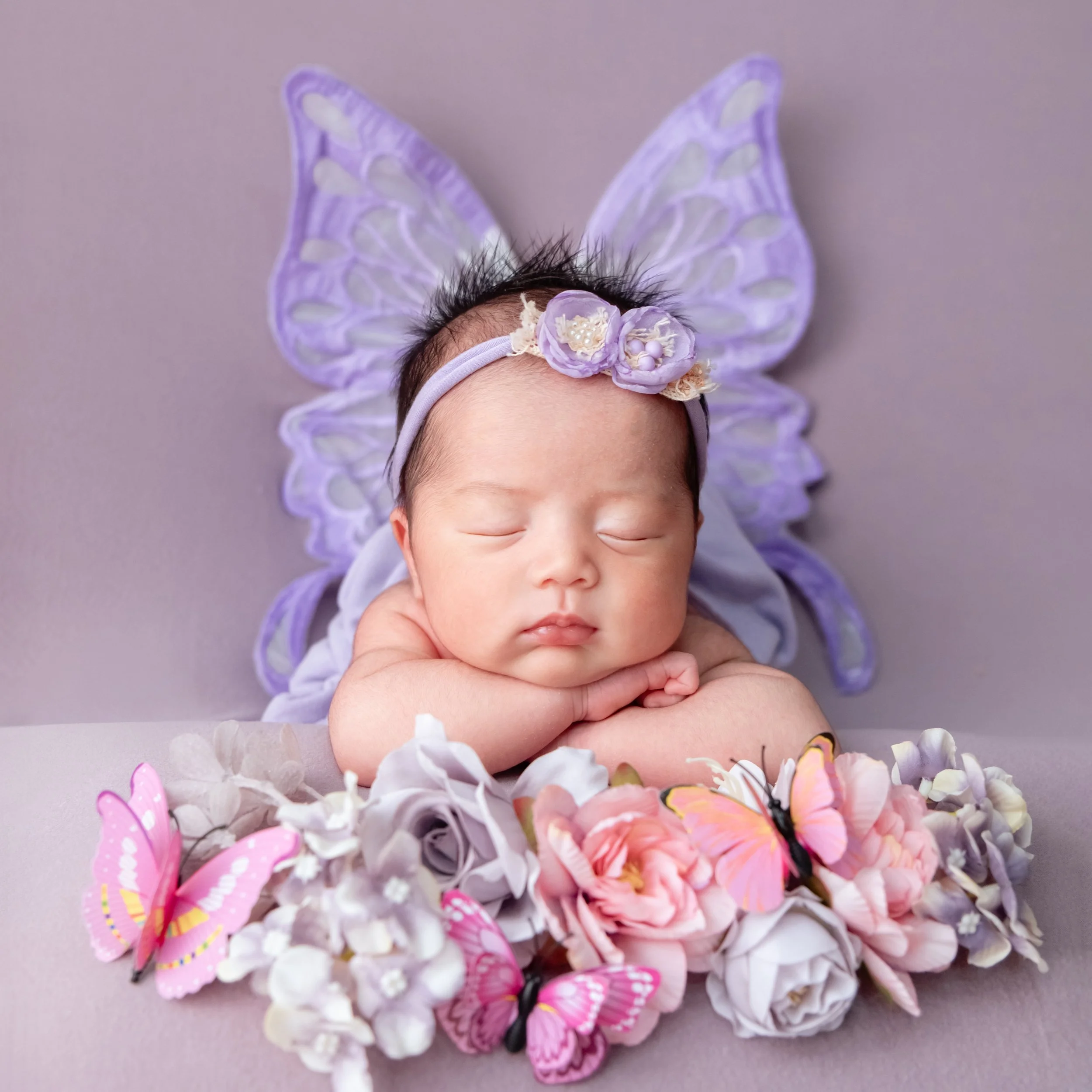 Sleeping newborn resting the chin on folded arms with purple butterfly wings behind and pink flowers in front