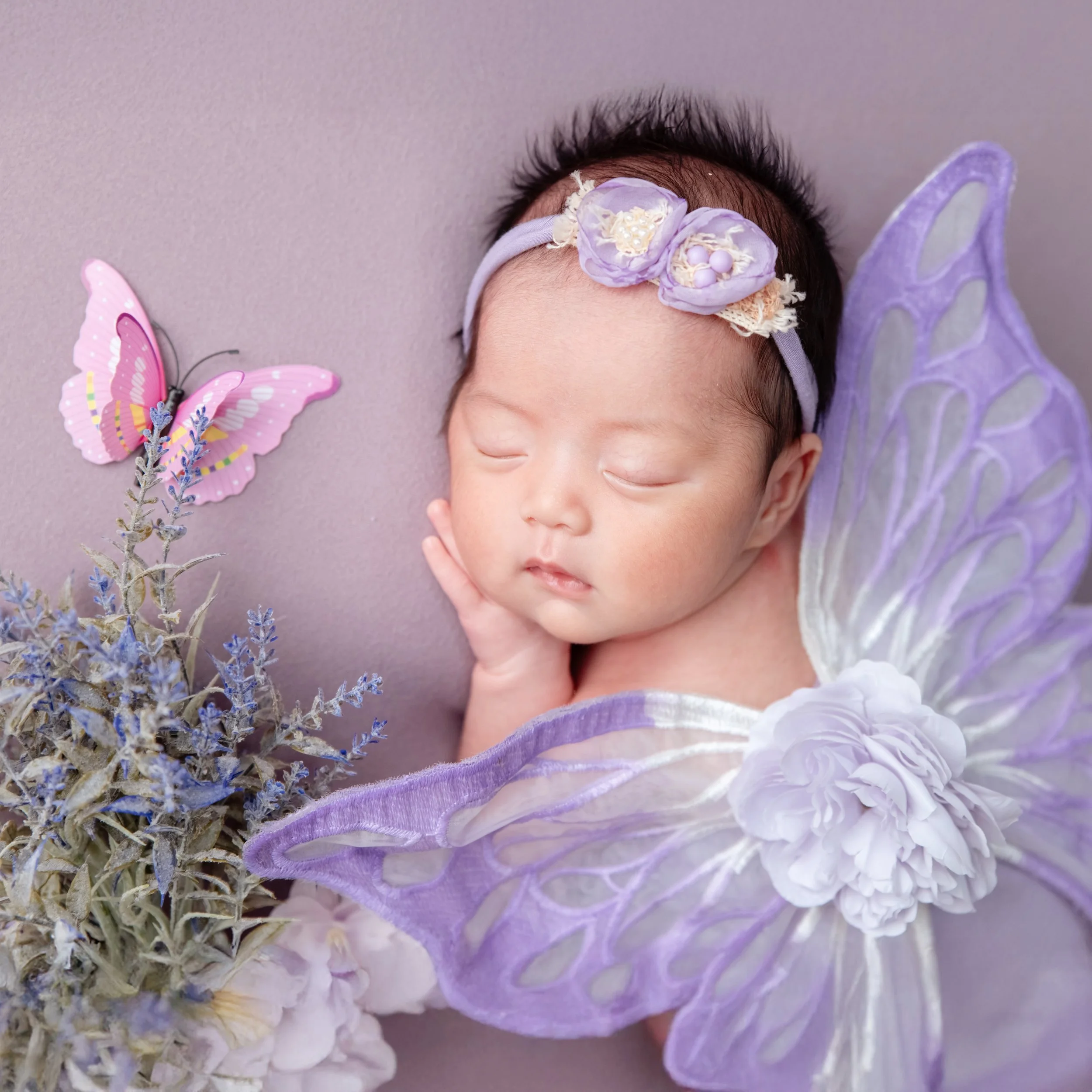 Sleeping newborn with a lavender floral headband and purple butterfly wings beside pastel flowers