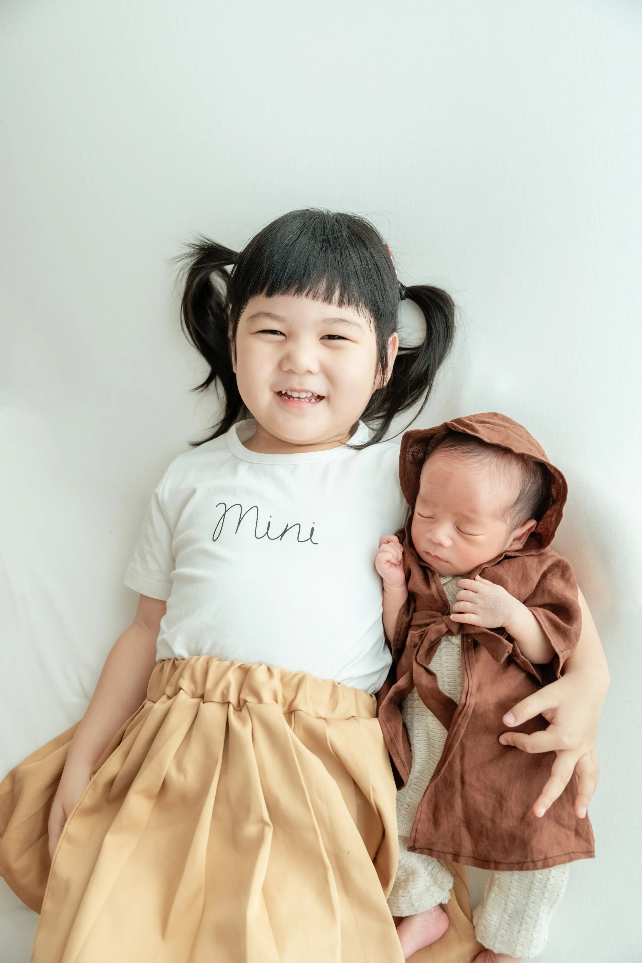 Smiling older sibling lying beside a sleeping newborn in a brown Jedi robe against a pale backdrop