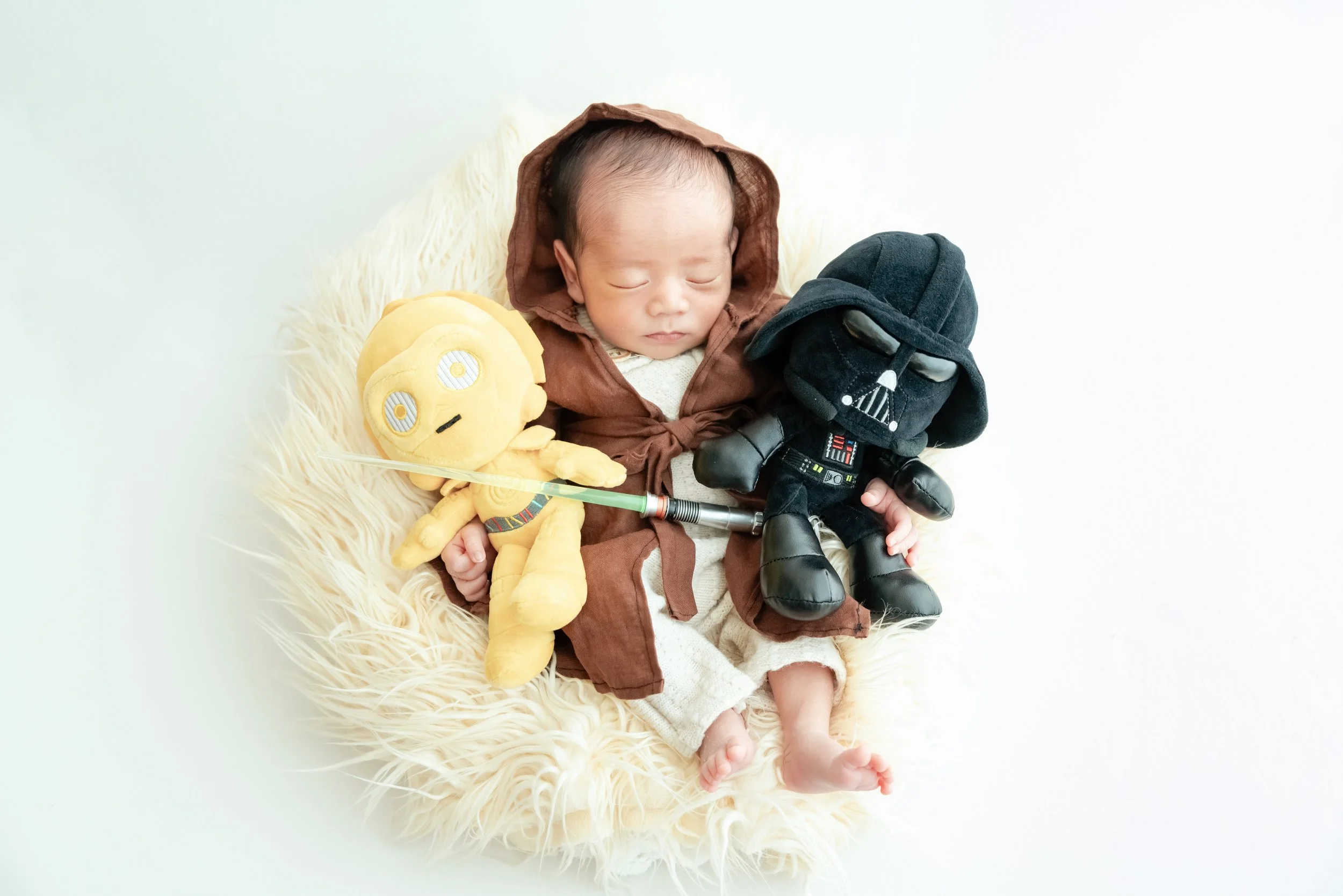 Sleeping newborn in a hooded Jedi robe nestled in a cream basket between C-3PO and Darth Vader plush toys
