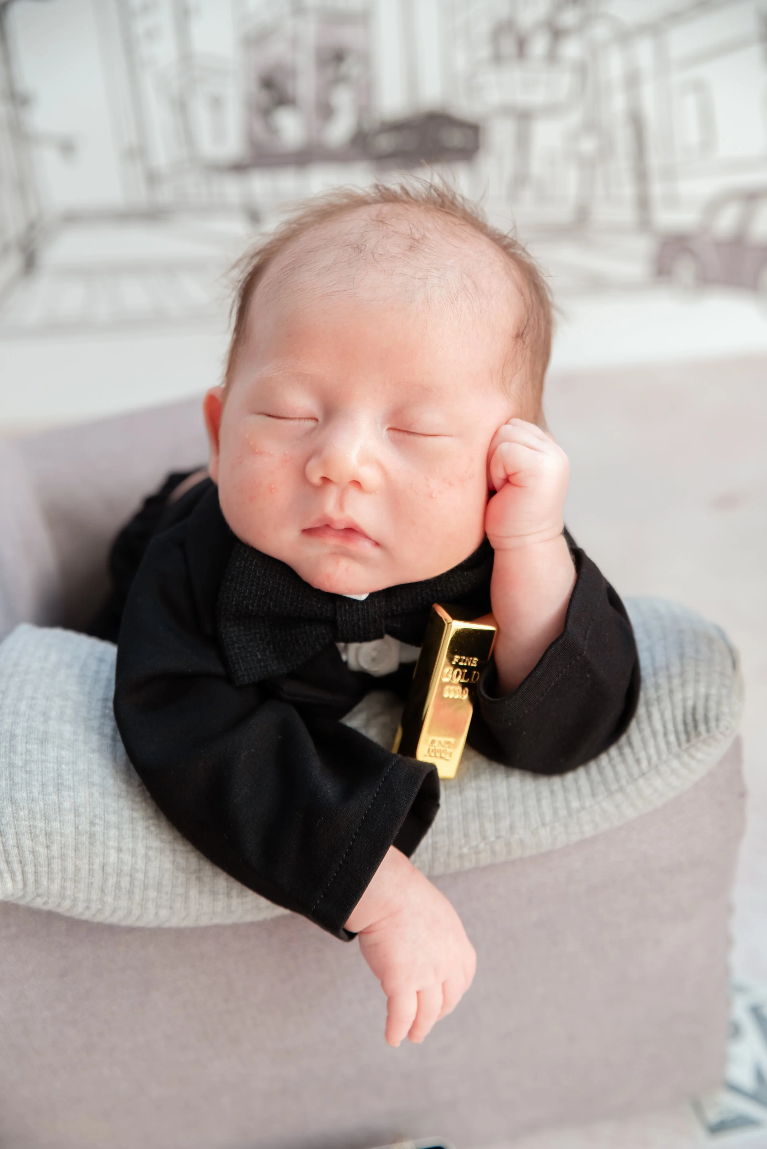 Close-up of a sleeping newborn in a black suit resting one hand beside the head with a toy gold bar on a grey armchair