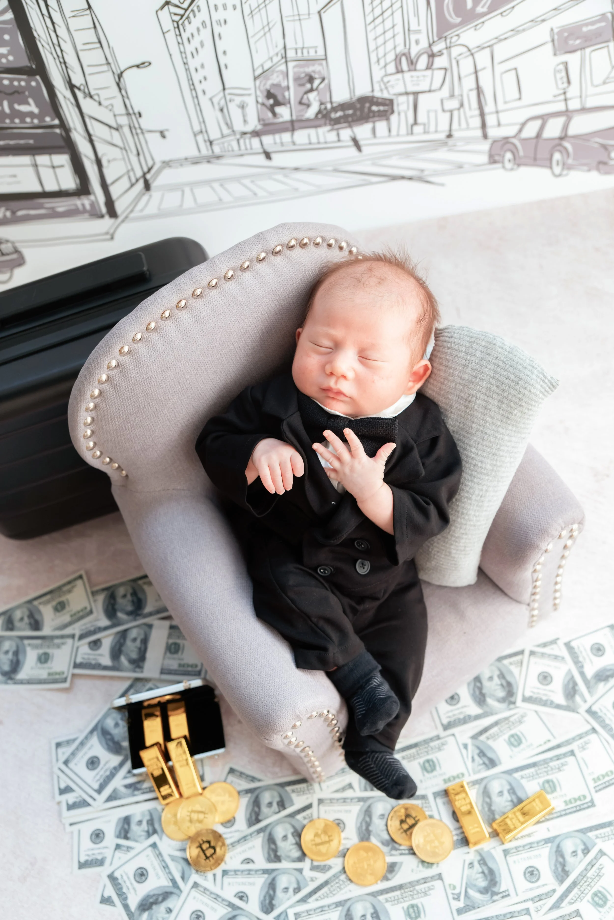 Sleeping newborn in a black suit seated in a miniature grey armchair surrounded by toy banknotes and gold bars