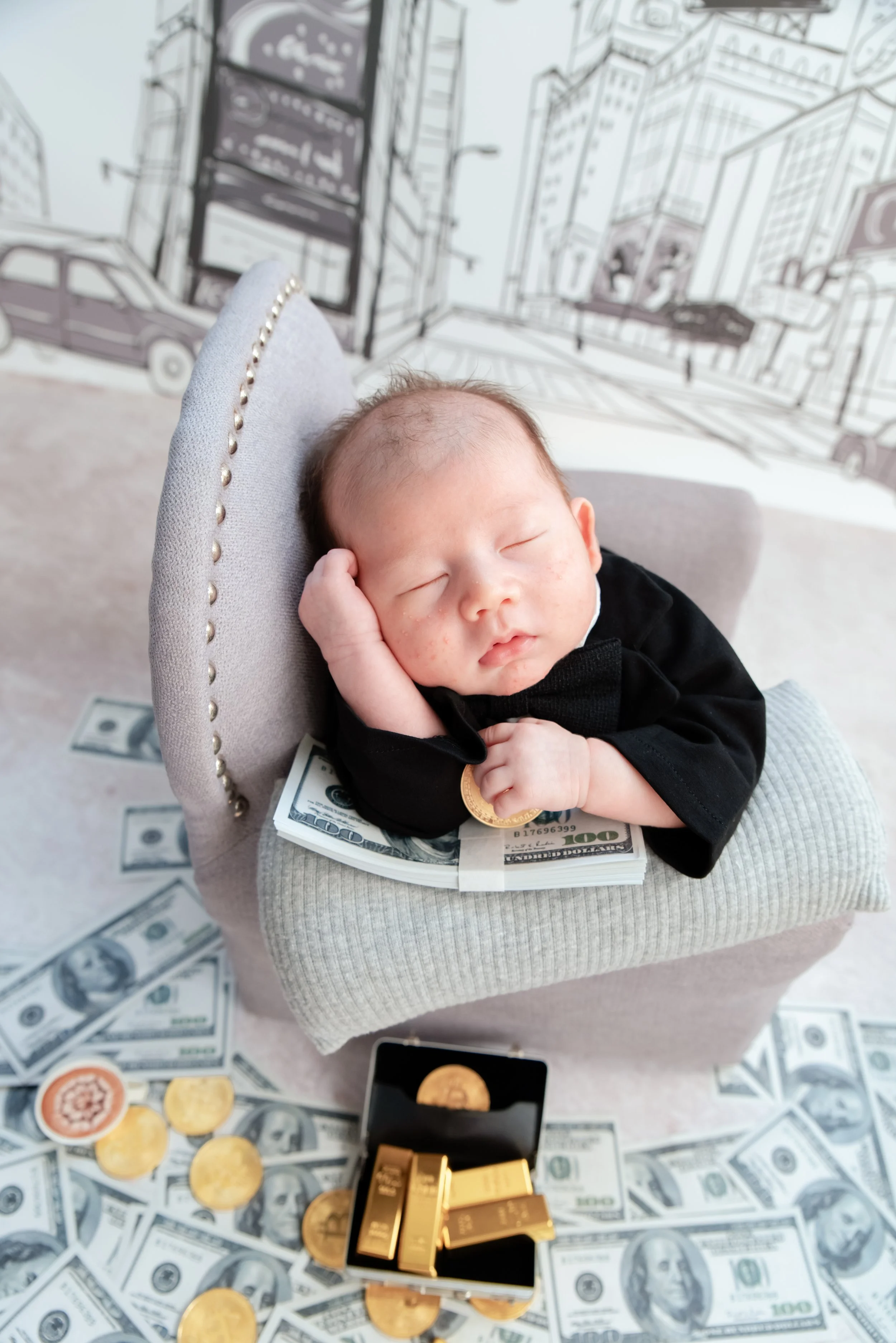 Sleeping newborn in a black suit leaning on a miniature grey armchair with hands beside the cheek and toy gold bars below