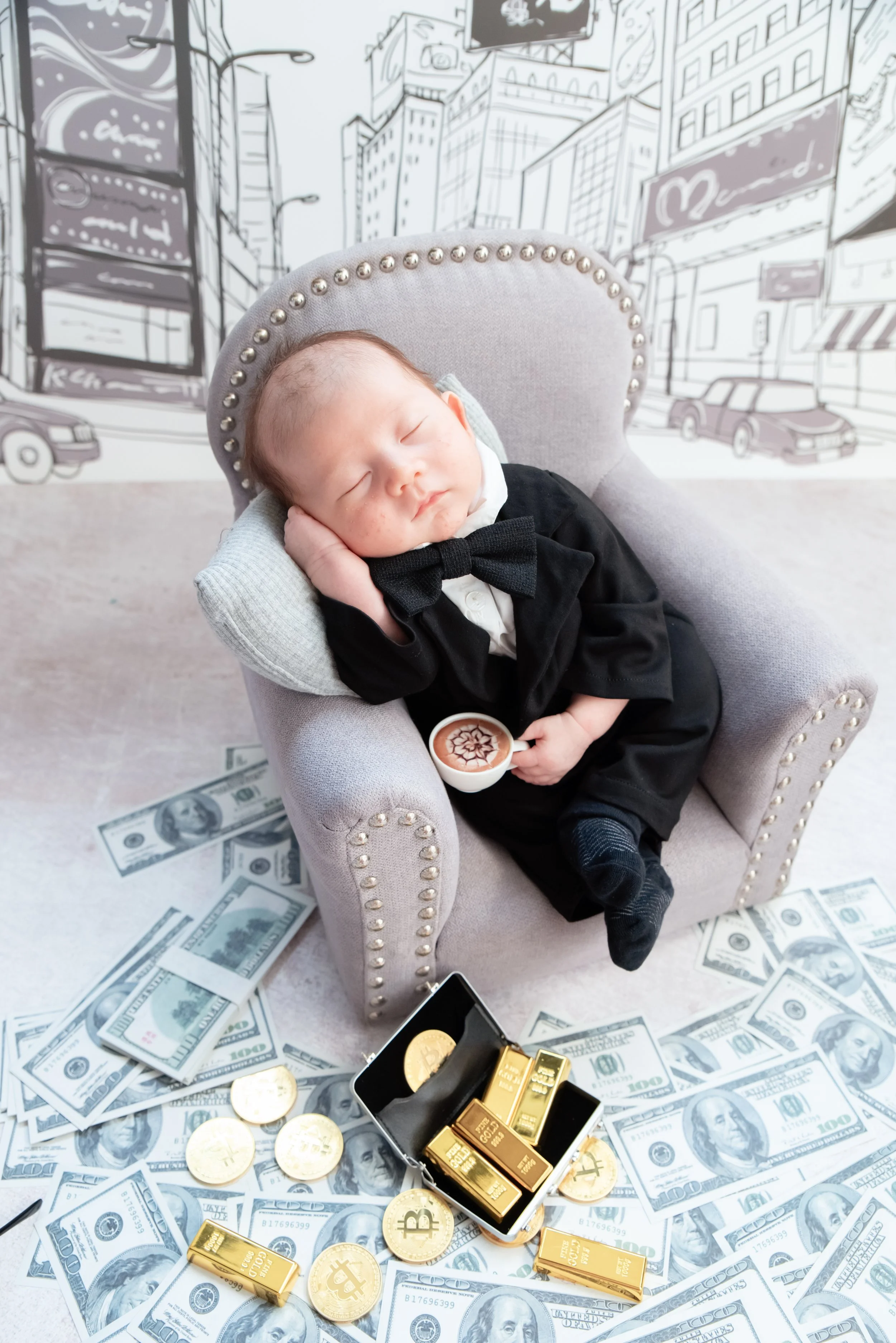 Sleeping newborn in a black suit reclining in a miniature grey armchair with one hand beside the cheek, toy banknotes and gold bars