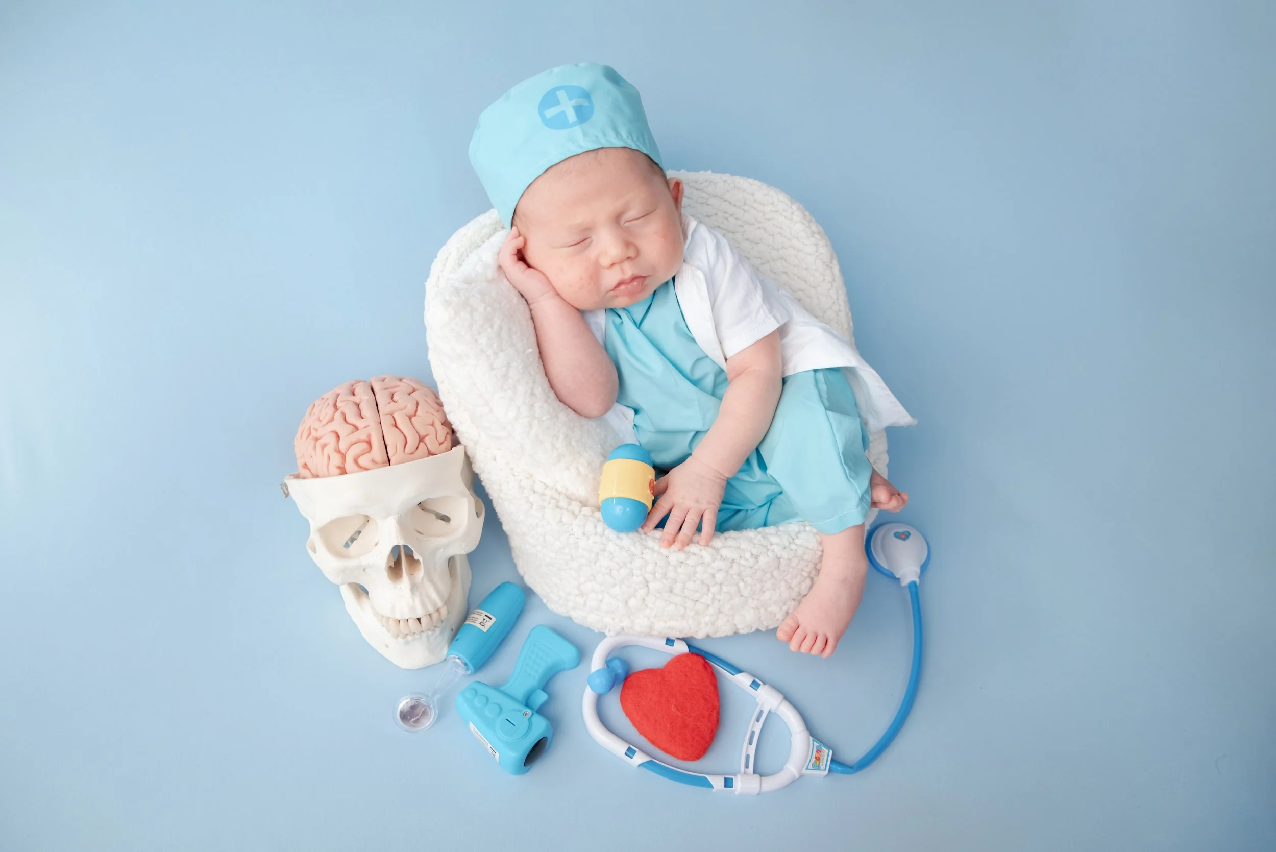 Sleeping newborn in blue medical scrubs and a white coat reclining in a fluffy basket surrounded by toy medical props