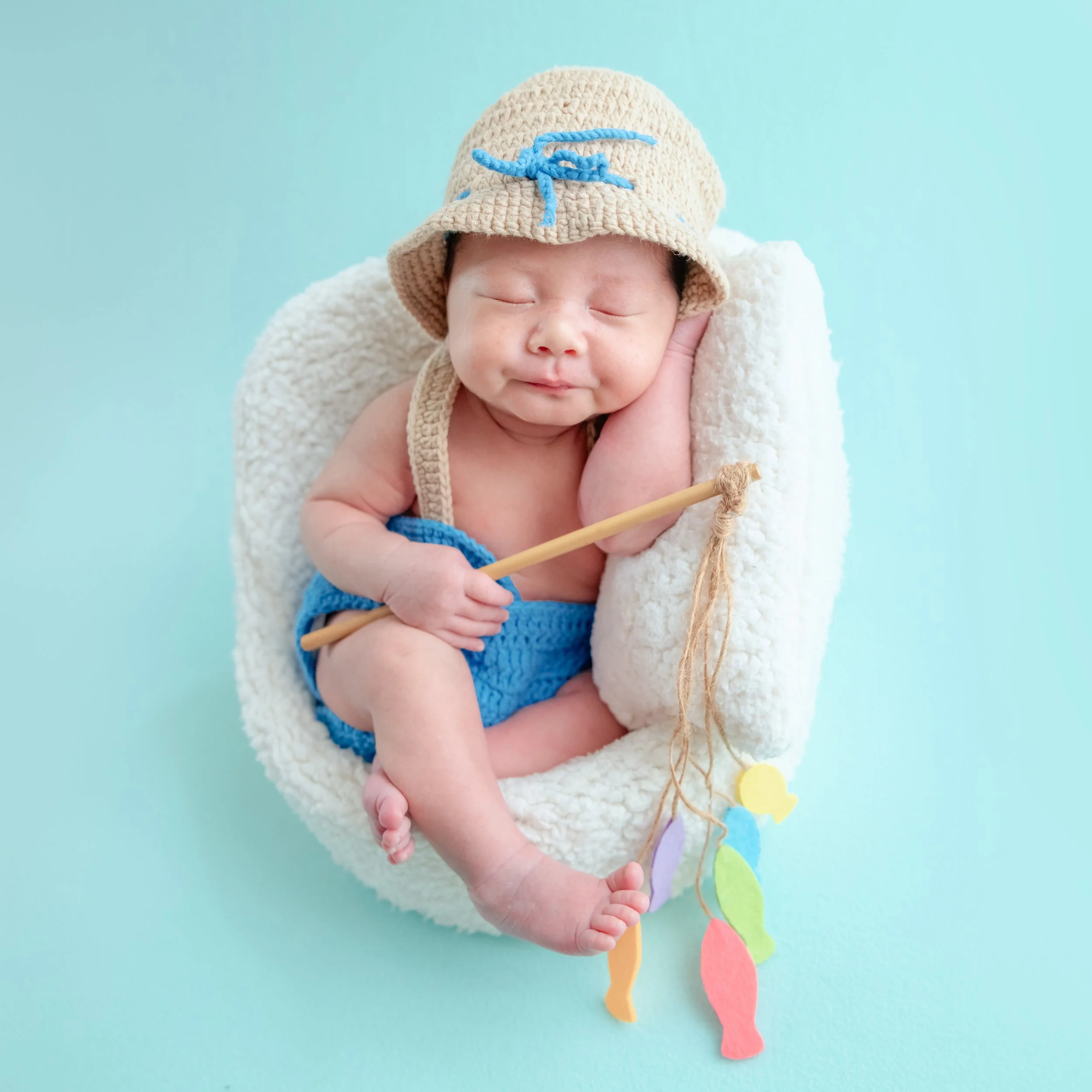 Sleeping newborn in a straw hat and blue knitted shorts sitting in a white basket with a toy fishing rod and hanging colourful fish