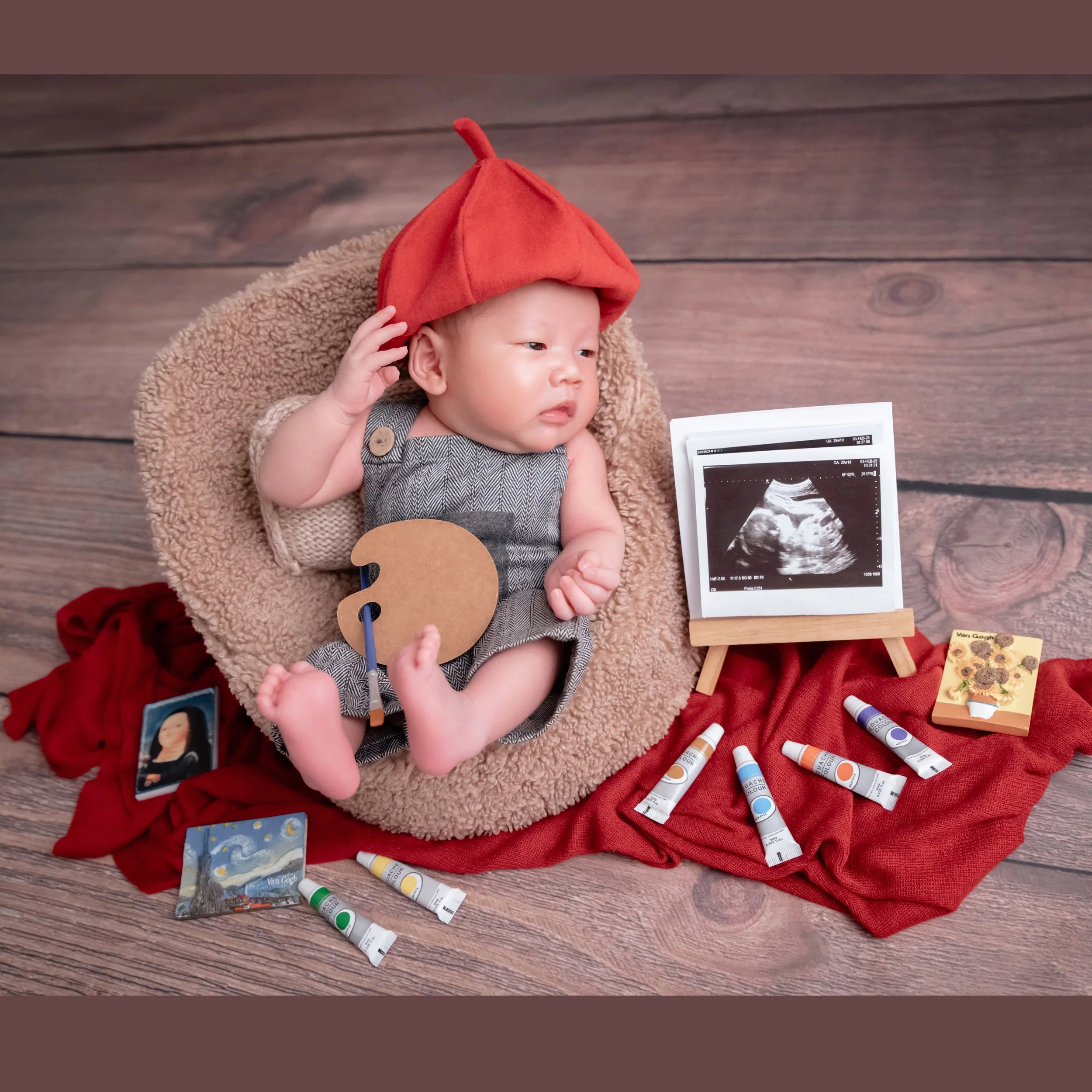 Awake newborn in a red painter beret and grey outfit holding a wooden palette in a beige basket beside brushes and paint tubes