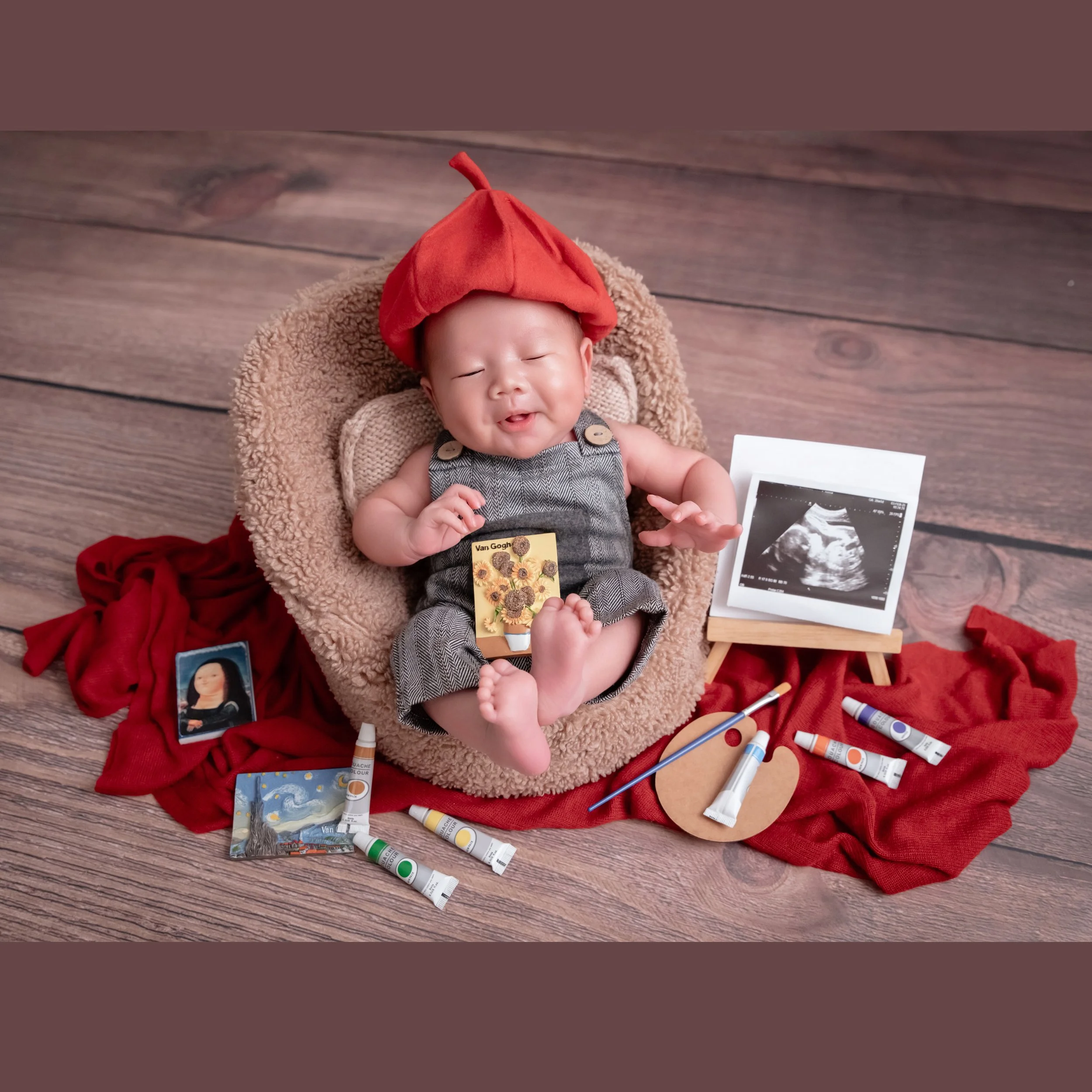 Yawning newborn wearing a red painter beret in a beige basket among paint tubes, brushes and miniature framed pictures