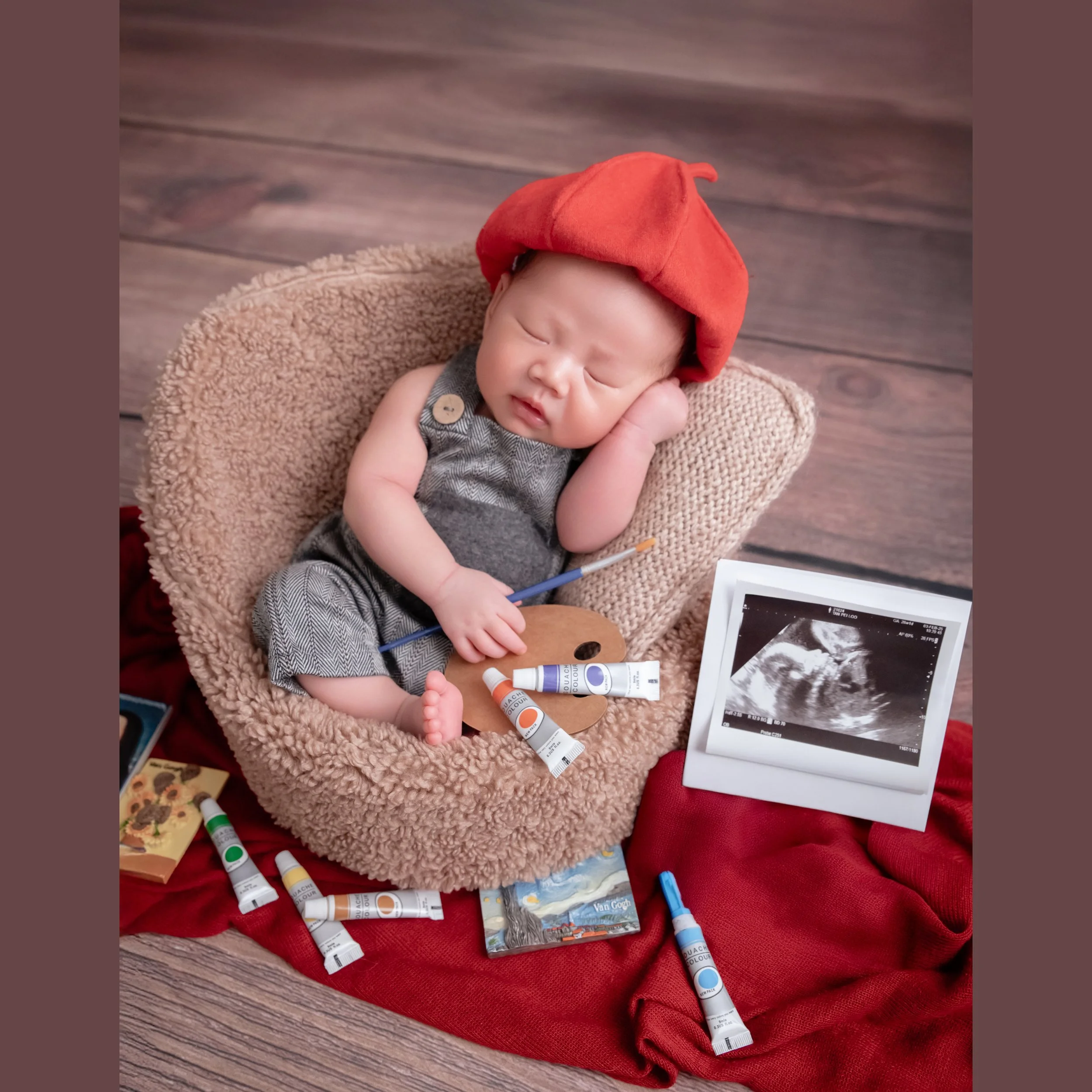 Sleeping newborn in a red painter beret and grey outfit with one hand beside the cheek in a beige basket among brushes and paint tubes