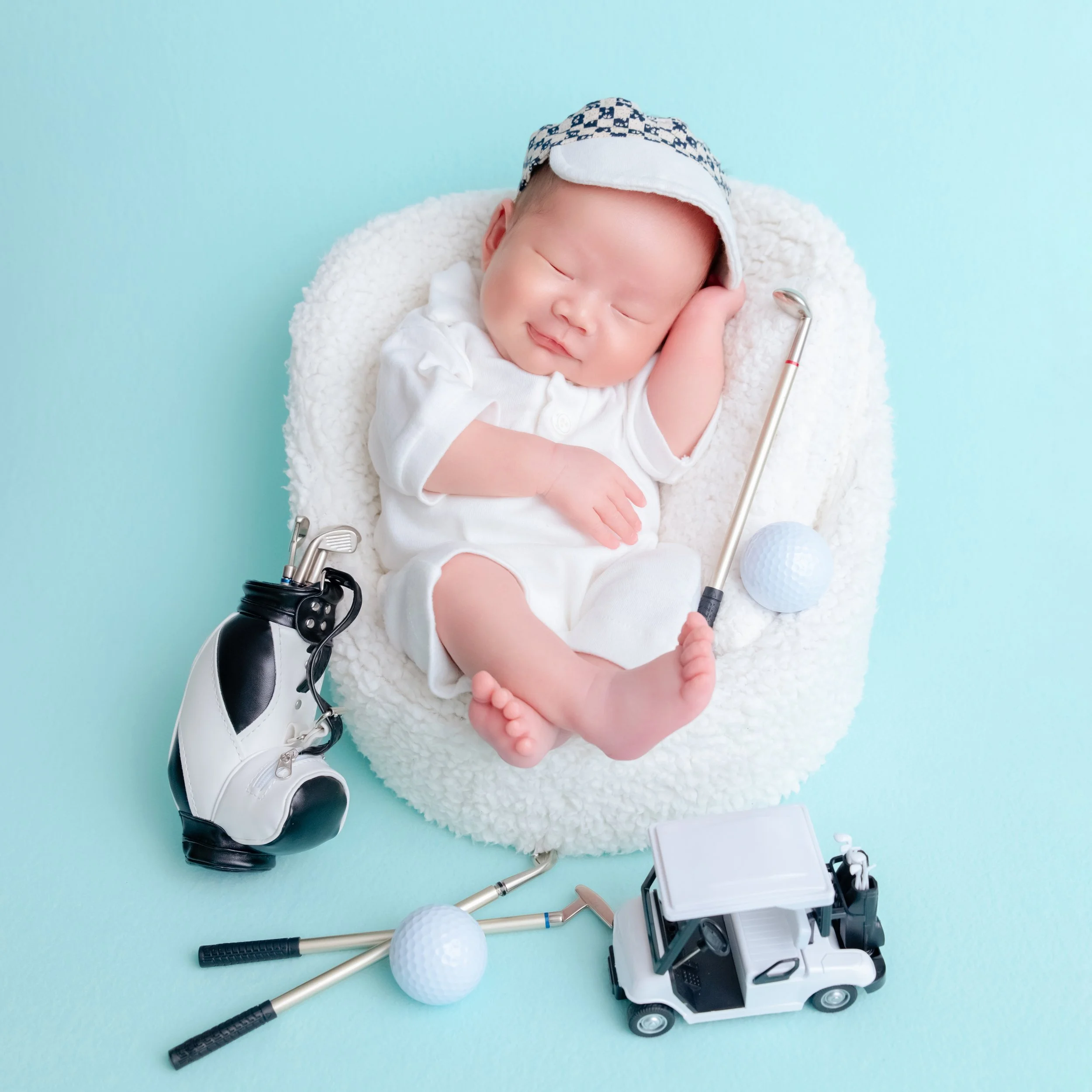 Sleeping newborn in a white golf outfit and checked visor reclining in a white basket with one hand behind the head beside a golf bag, clubs and toy golf cart