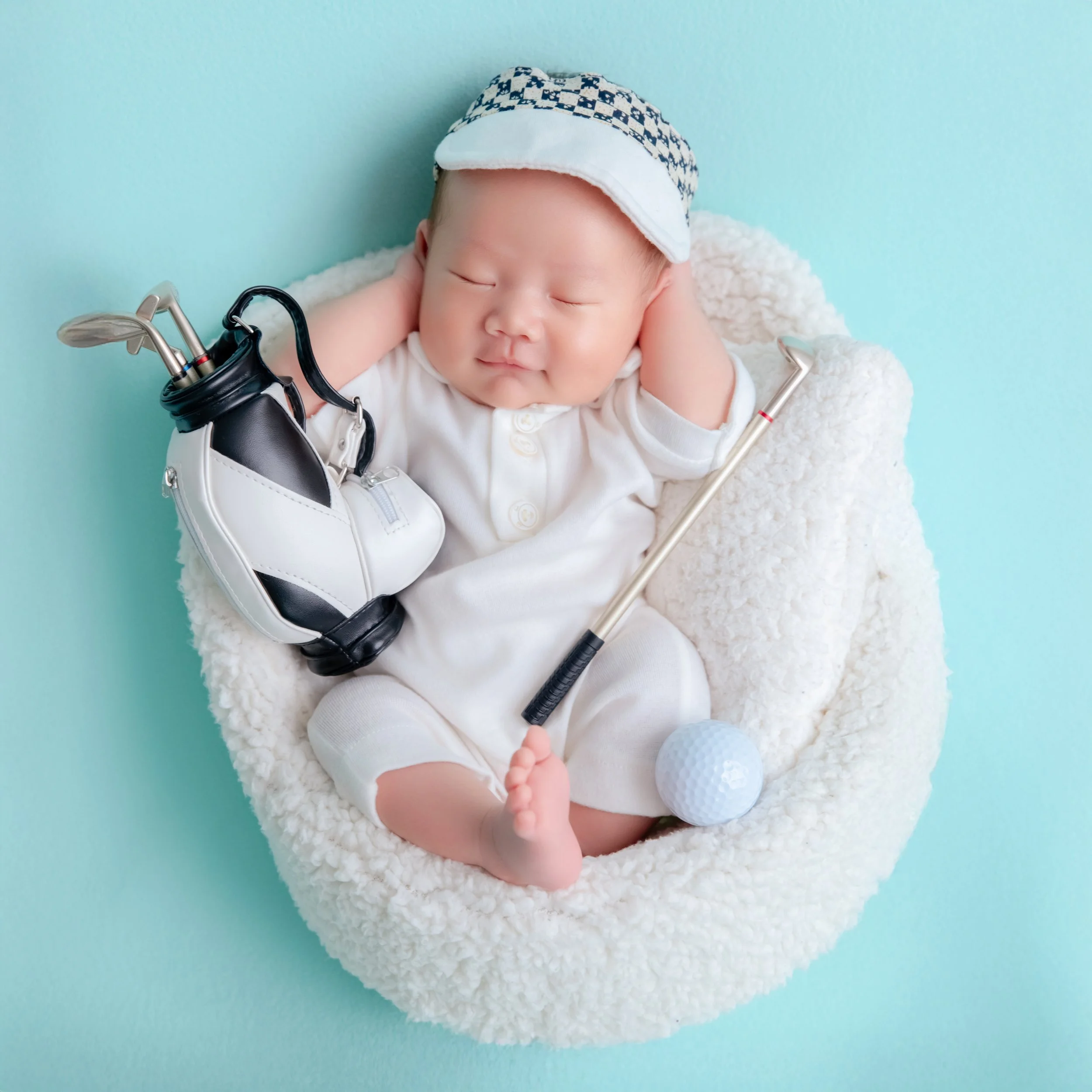 Yawning newborn in a white golf outfit and checked visor reclining in a white basket beside a miniature golf bag and clubs