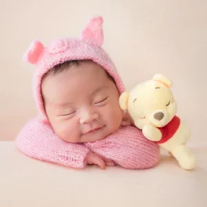 Smiling newborn in a pink Piglet hat resting on folded arms beside a Winnie the Pooh plush on a cream backdrop