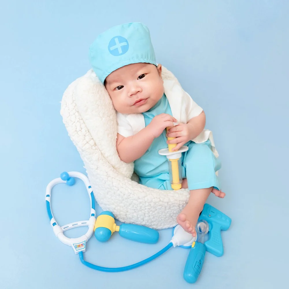 Awake baby in a blue medical cap and white coat holding a yellow toy instrument in a fluffy basket beside a stethoscope