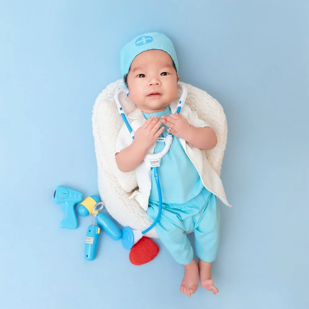Awake baby in blue medical scrubs and a white coat lying in a fluffy basket with hands together beside toy medical instruments