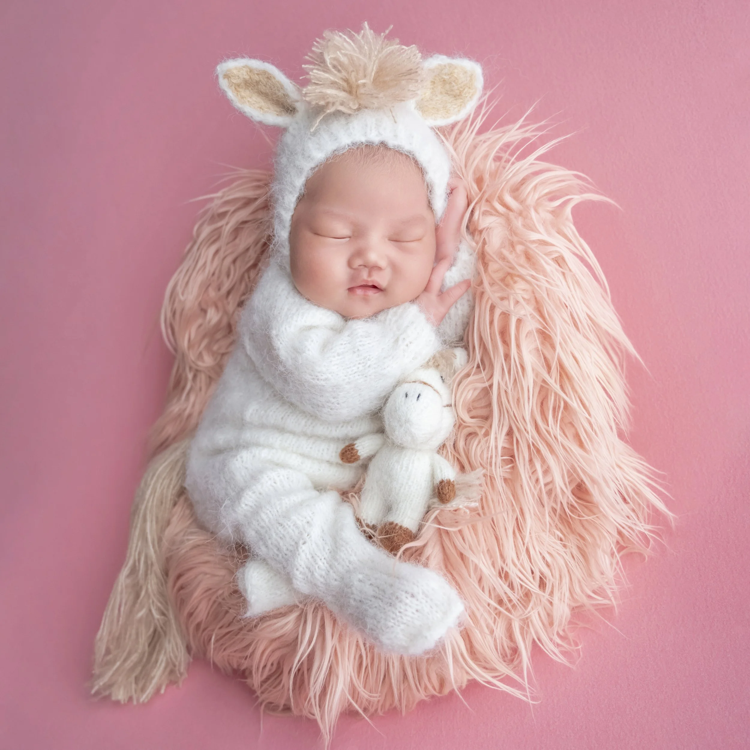 Sleeping newborn in a white knitted horse outfit with one hand beside the cheek and a horse toy on pink fur