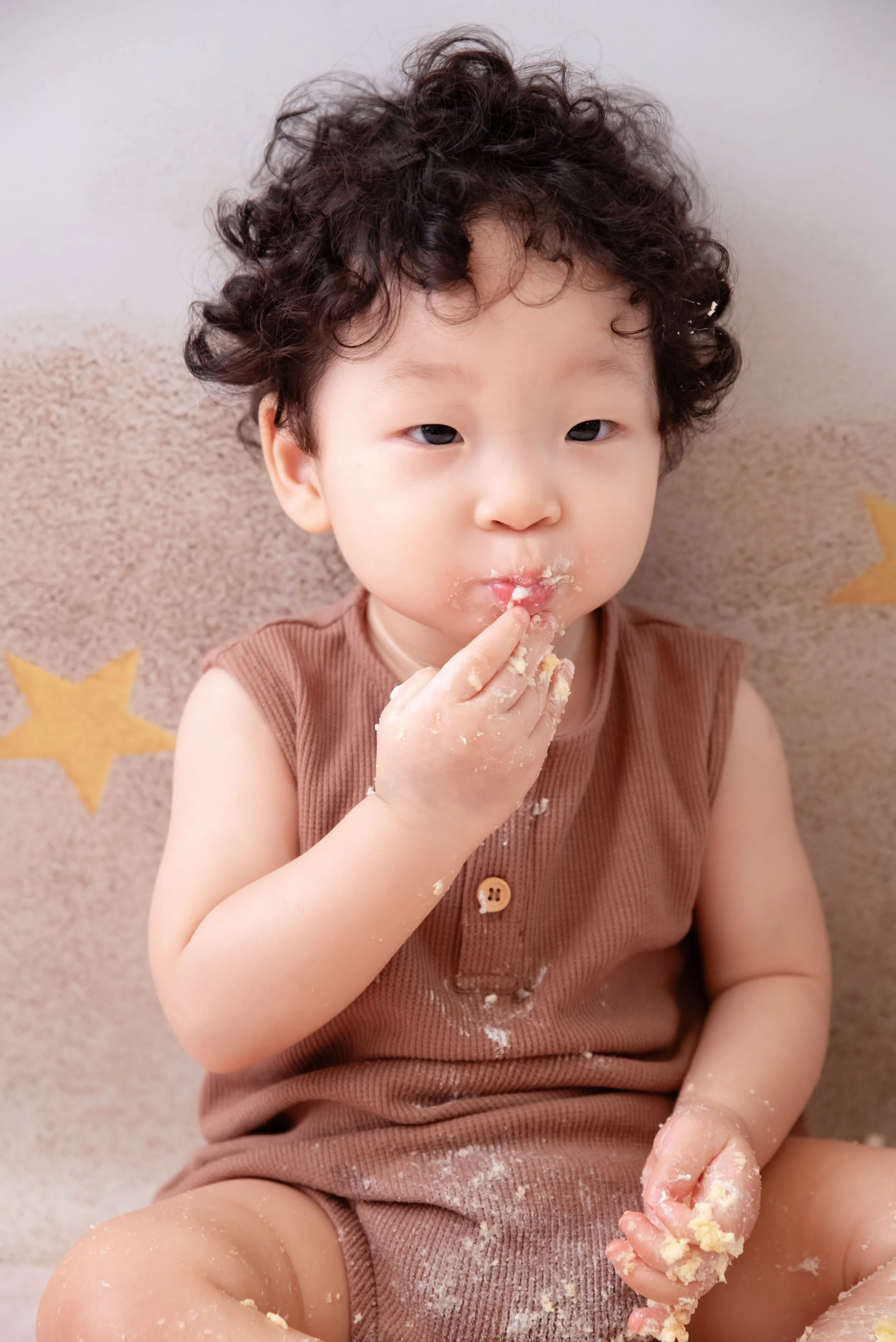 Curly-haired baby in a brown romper tasting frosting with cake-covered fingers