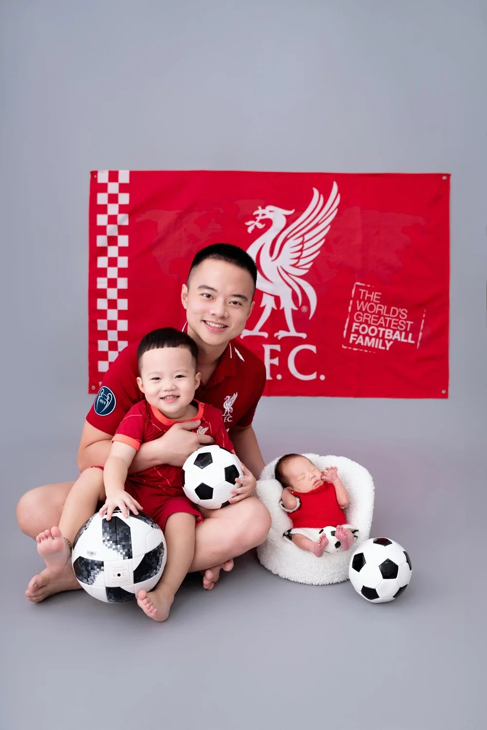 Smiling father and older son wearing red football shirts beside a sleeping newborn in a matching red outfit, photographed by Mamamiyo Photography in Singapore