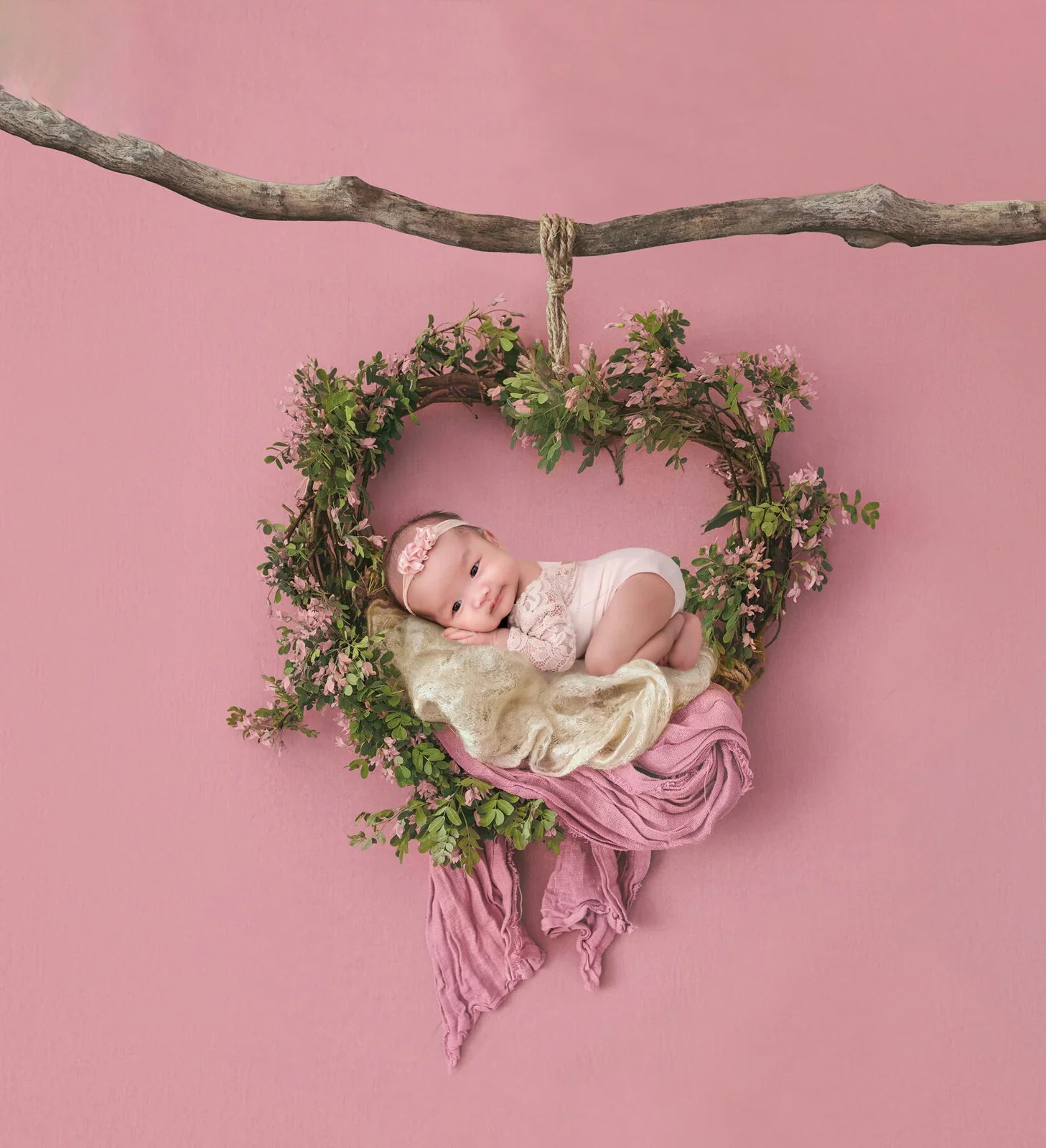 Newborn resting in a heart-shaped hanging floral nest against a pink background in a digital composition