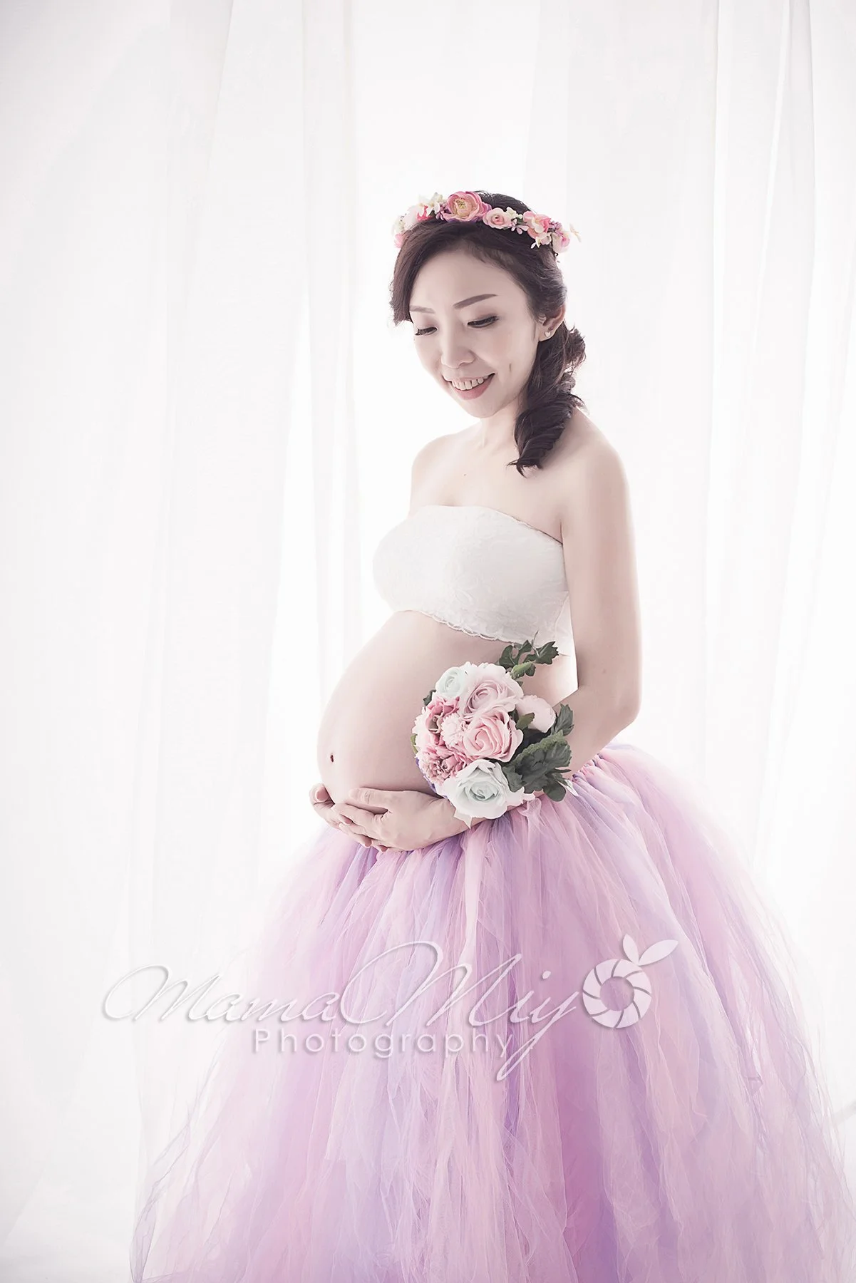Pregnant woman in a pink and lilac tutu skirt with a flower crown, holding pink roses beside white curtains