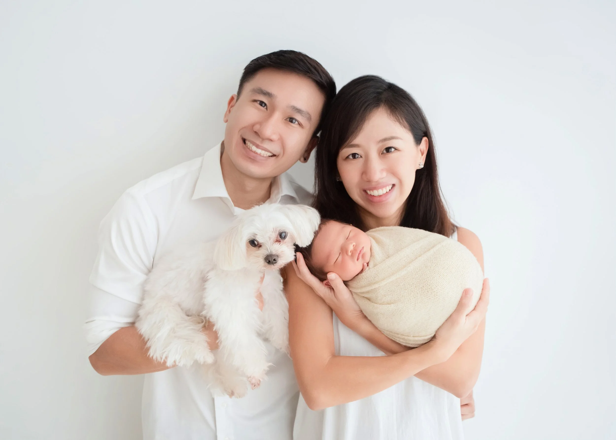 Newborn and family photography with pet by Mamamiyo Photography, Singapore: smiling parents holding their sleeping baby beside their white dog.