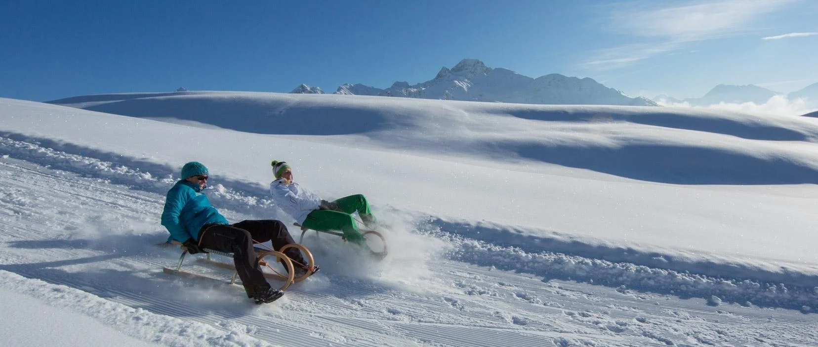 Zwei Personen rodeln auf einem schneebedeckten Hang bei sonnigem Wetter, im Hintergrund Berge.