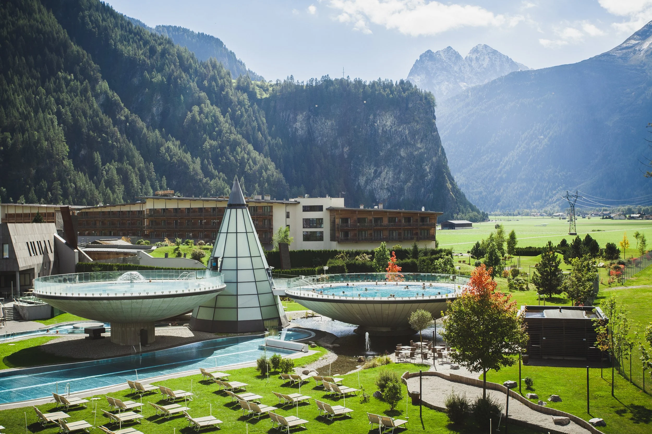 Mountainresort mit futuristischen Poolanlagen, umgeben von Wiesen und Bergen, im Hintergrund Holzhotelgebäude, Bäume und Liegestühle im Vordergrund.