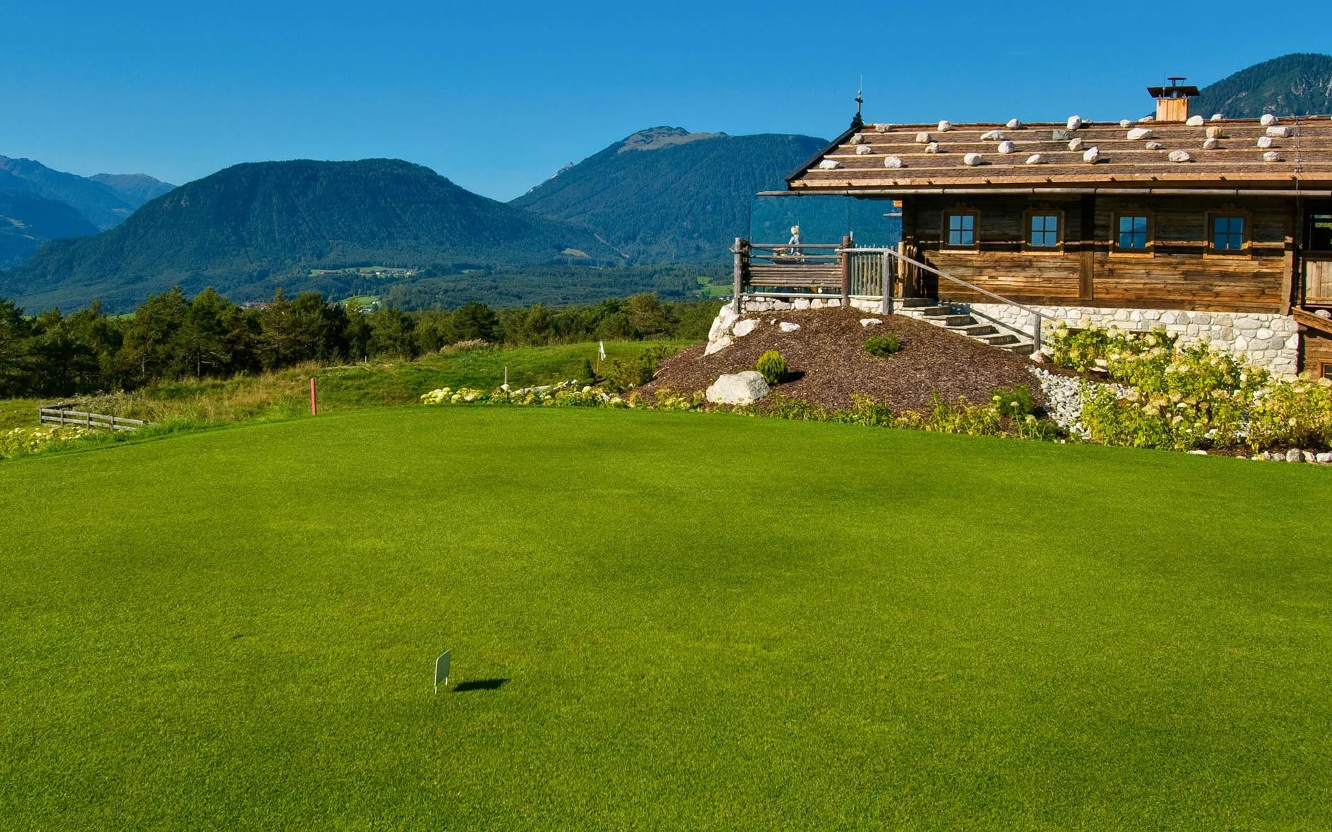 Grüner Golfplatz vor einem Holzhaus mit Bergblick, blauer Himmel, bei Sonnenschein.