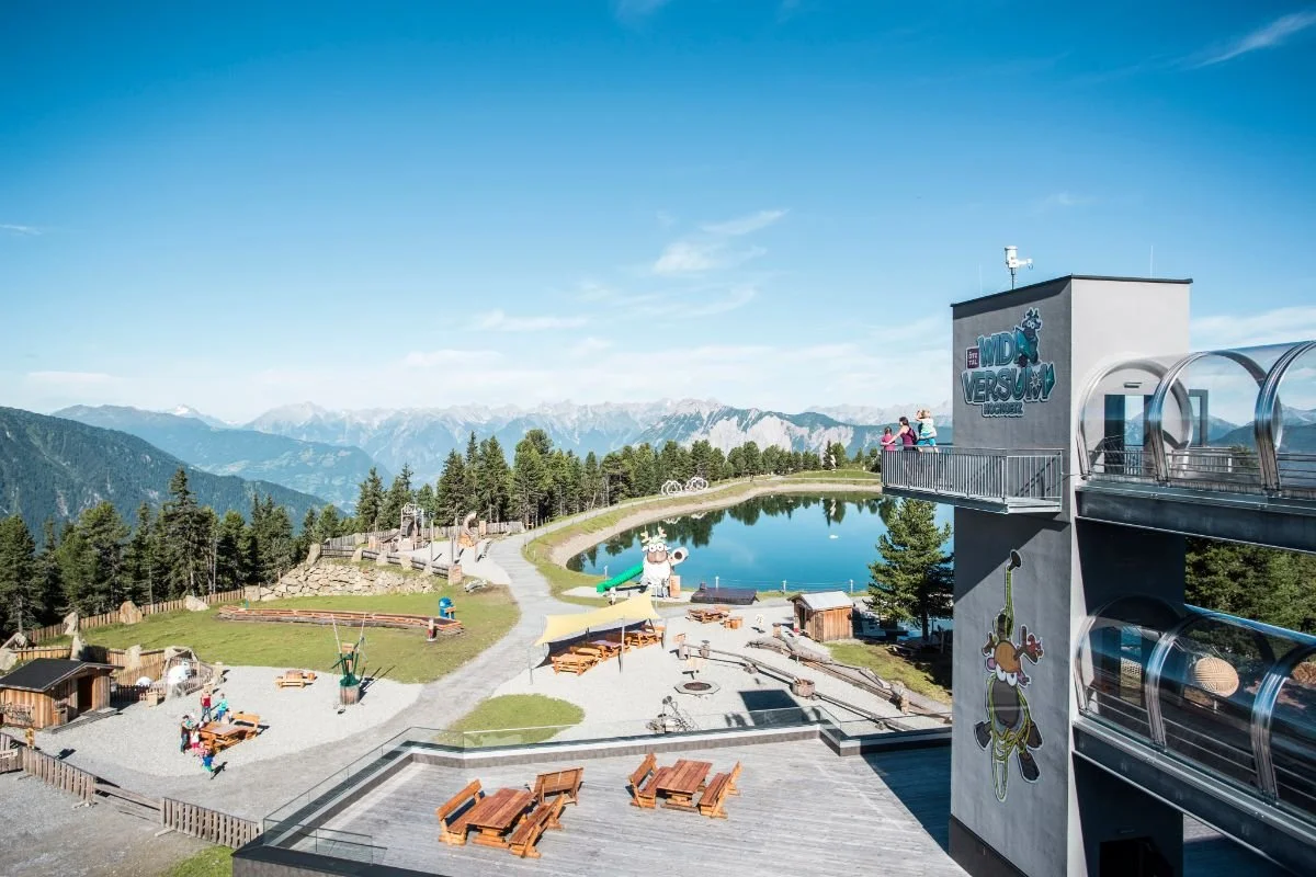 Berglandschaft mit einem Freizeitpark, einem künstlichen See und Spielplatz; Gebäudestruktur mit Aufschrift 'WIDIVERSEUM', umgeben von Wald und Bergpanorama. Kinder und Erwachsene auf einem Balkon.