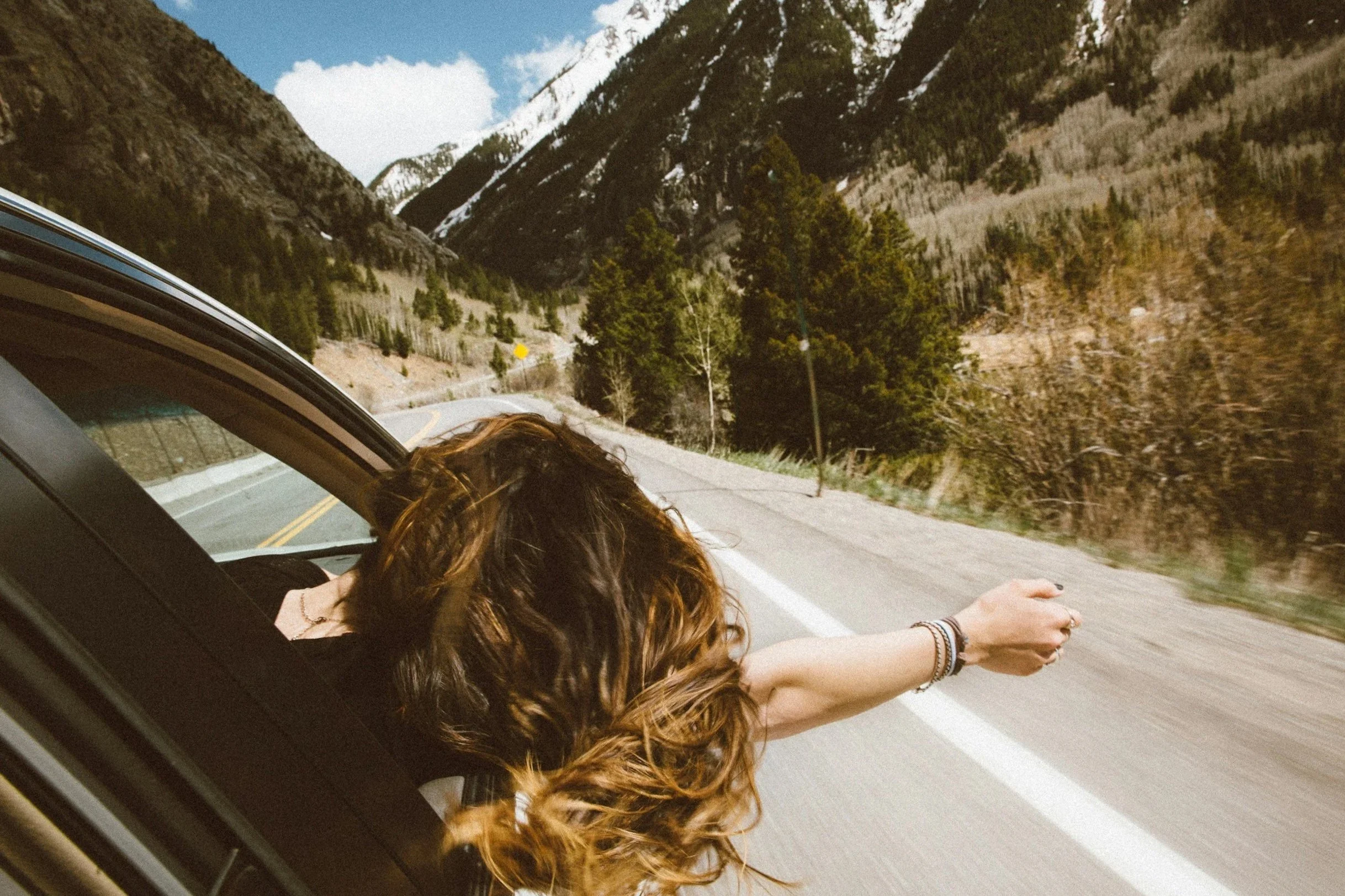 A person with brown, curly hair leaning out of a moving car window, reaching their arm out toward the landscape, with a mountainous backdrop and a winding road.
