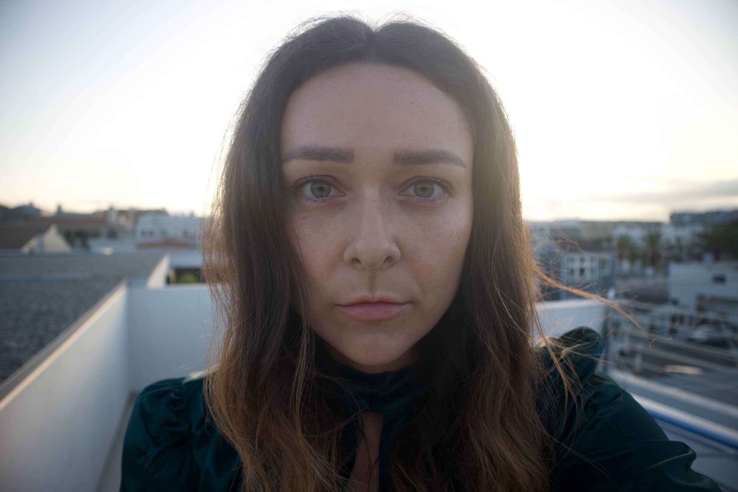 A close-up photo of a woman with long brown hair and blue eyes, standing outdoors on a rooftop during sunset, with buildings and a cloudy sky in the background.