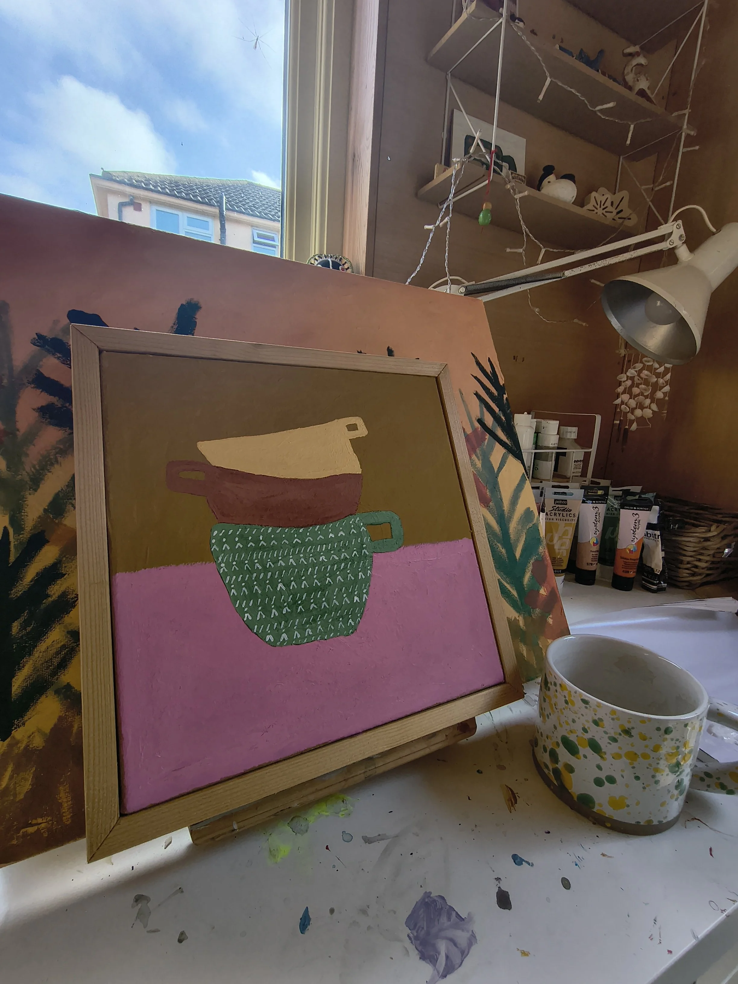A pink painting of a decorative teapot with a green patterned bottom and a light brown top. The painting is on an easel in a cluttered art studio with paint tubes, a large patterned mug, and shelves with figurines and art supplies.