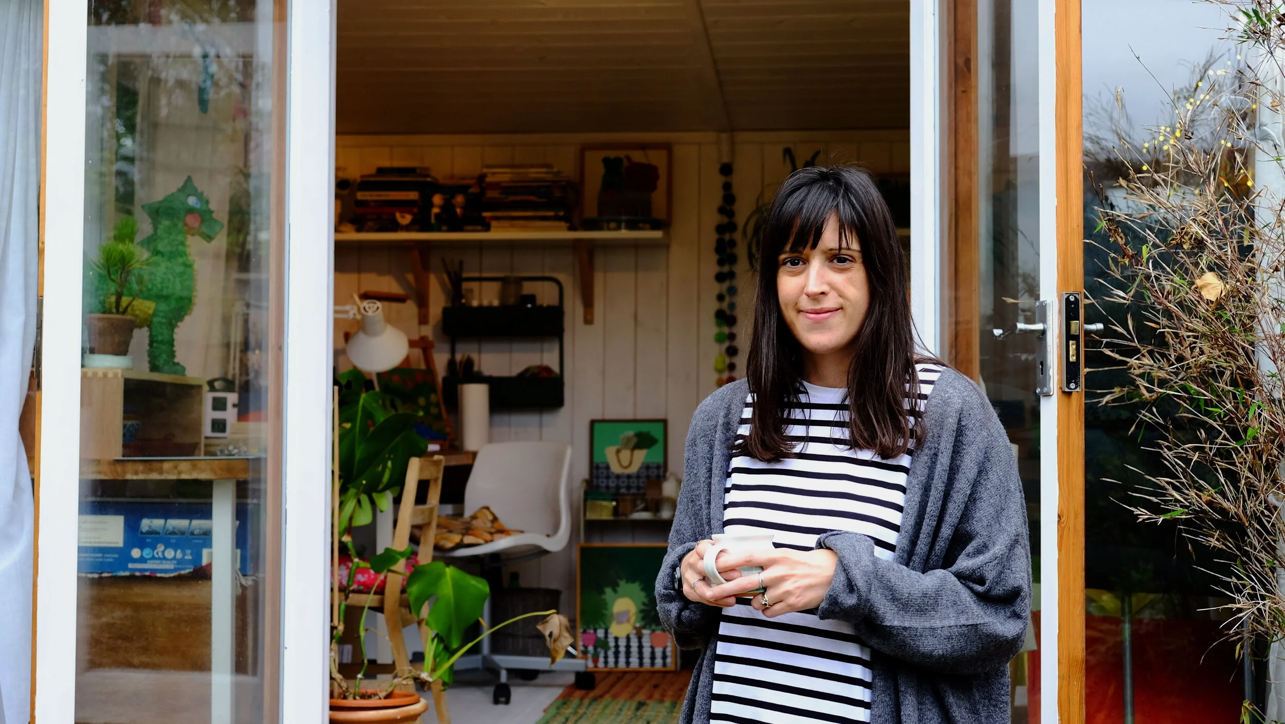 A young woman with dark hair and a striped shirt standing outside a screened-in porch, holding a mug, with plants and furniture visible inside.