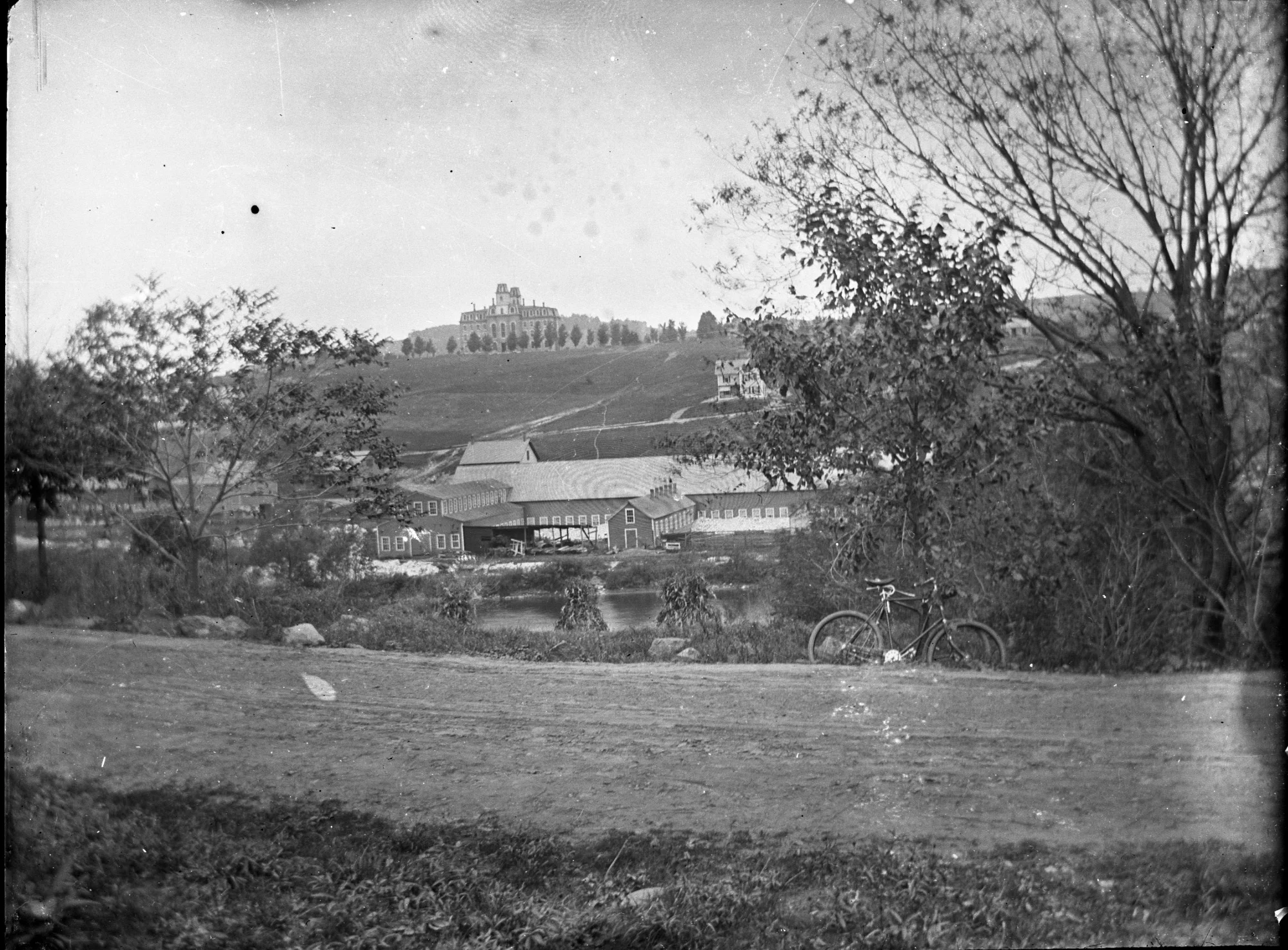 Bicycle and Granite Sheds - A bicycle rests in a ditch along Berlin Street, Montpelier, Vermont. Across the Winooski River is a granite shed and on top of the hill is Vermont Methodist Seminary. The Vermont Methodist Seminary is now the Vermont College of Fine Arts (VCFA). Courtesy the Vermont Historical Society.