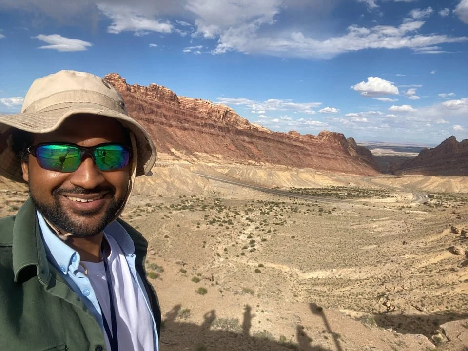 Paleontologist Cassius Morrison in front of a desert landscape, in tans and reddish stone, showing exposures of the Morrison Formation.